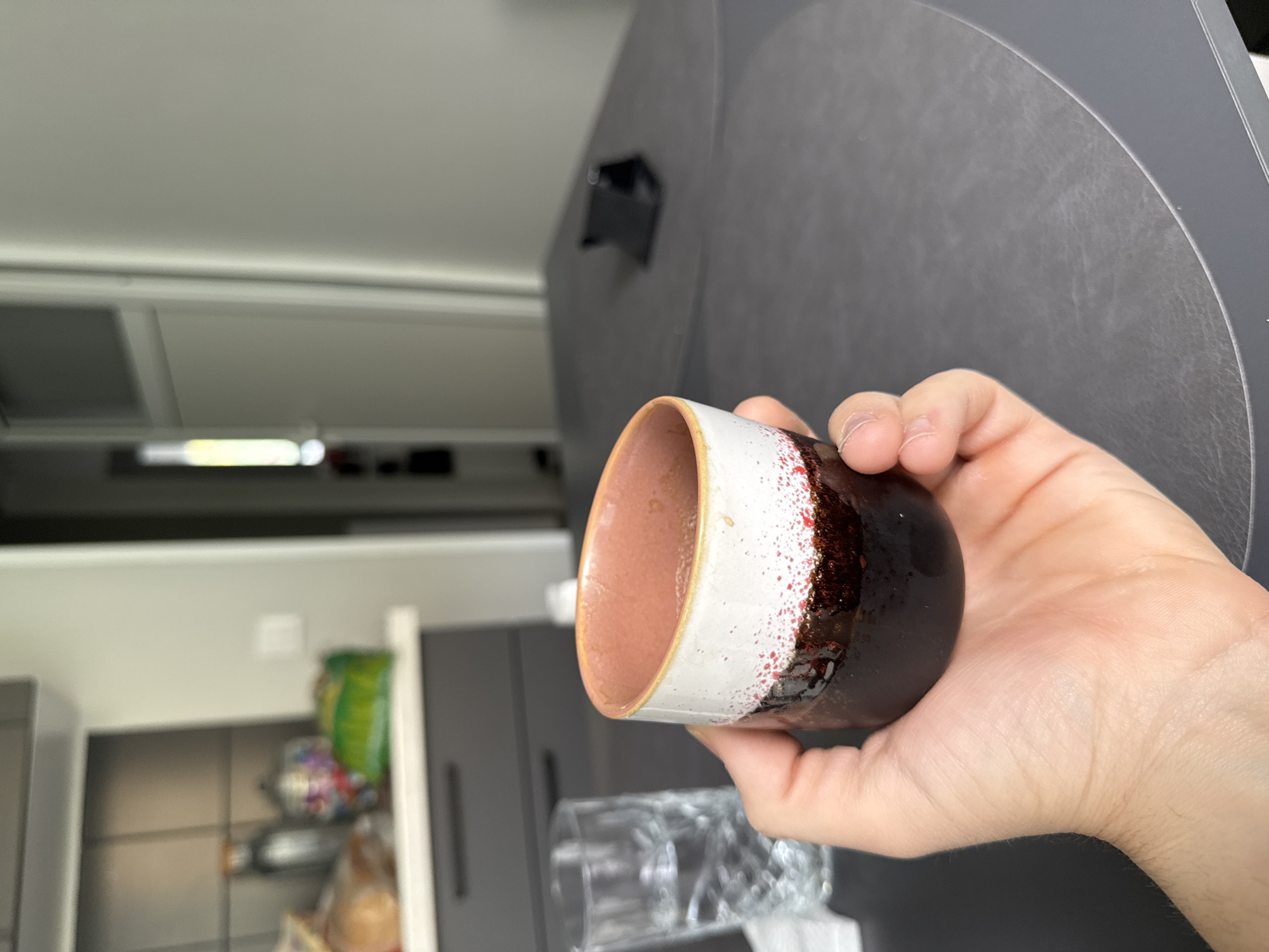 The image depicts a person's hand holding a cylindrical ceramic cup with a brown interior, a white band near the top, and a dark brown exterior with red speckles. The background shows a kitchen setting with grey cabinets and drawers. A set of clear glasses are visible on the countertop. There's a white fluorescent light on the ceiling. Based on the information available, there is nothing to conclude that the photo was taken in Stara Zagora, Bulgaria.