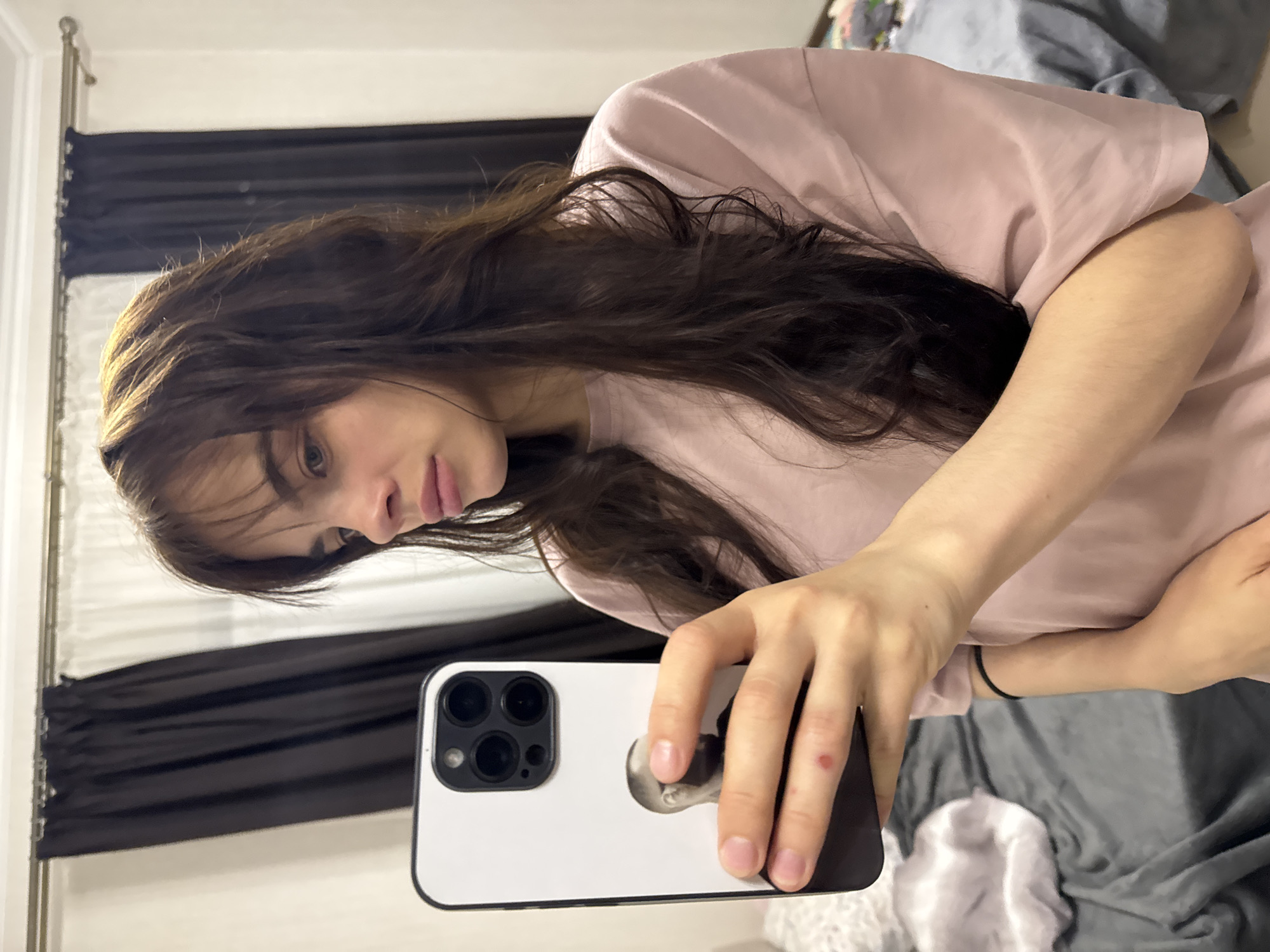 A young woman takes a mirror selfie in what appears to be a bedroom setting in Rostov-on-Don, Russia. The image is presented rotated 90 degrees counter-clockwise. She has long, dark brown hair that cascades over her shoulders and partially across her face. She is wearing a light pink, short-sleeved t-shirt. Her expression is neutral, and she appears to be looking down towards the phone she is holding.
In the foreground, her right hand holds a white smartphone, identifiable as an iPhone due to its distinctive three-lens camera module. The phone is encased in a white cover that features a black and white graphic, depicting what looks like a stylized human head or face. A small red mark is visible on the knuckle of her right index finger.
The background reveals dark curtains, likely on a window, behind her left side. To her right, a bed with gray bedding is partially visible. A gold-colored metallic frame, possibly part of a bedpost or headboard, can be seen on the far left of the reflection. The lighting suggests an indoor environment. There is no visible text in the image.