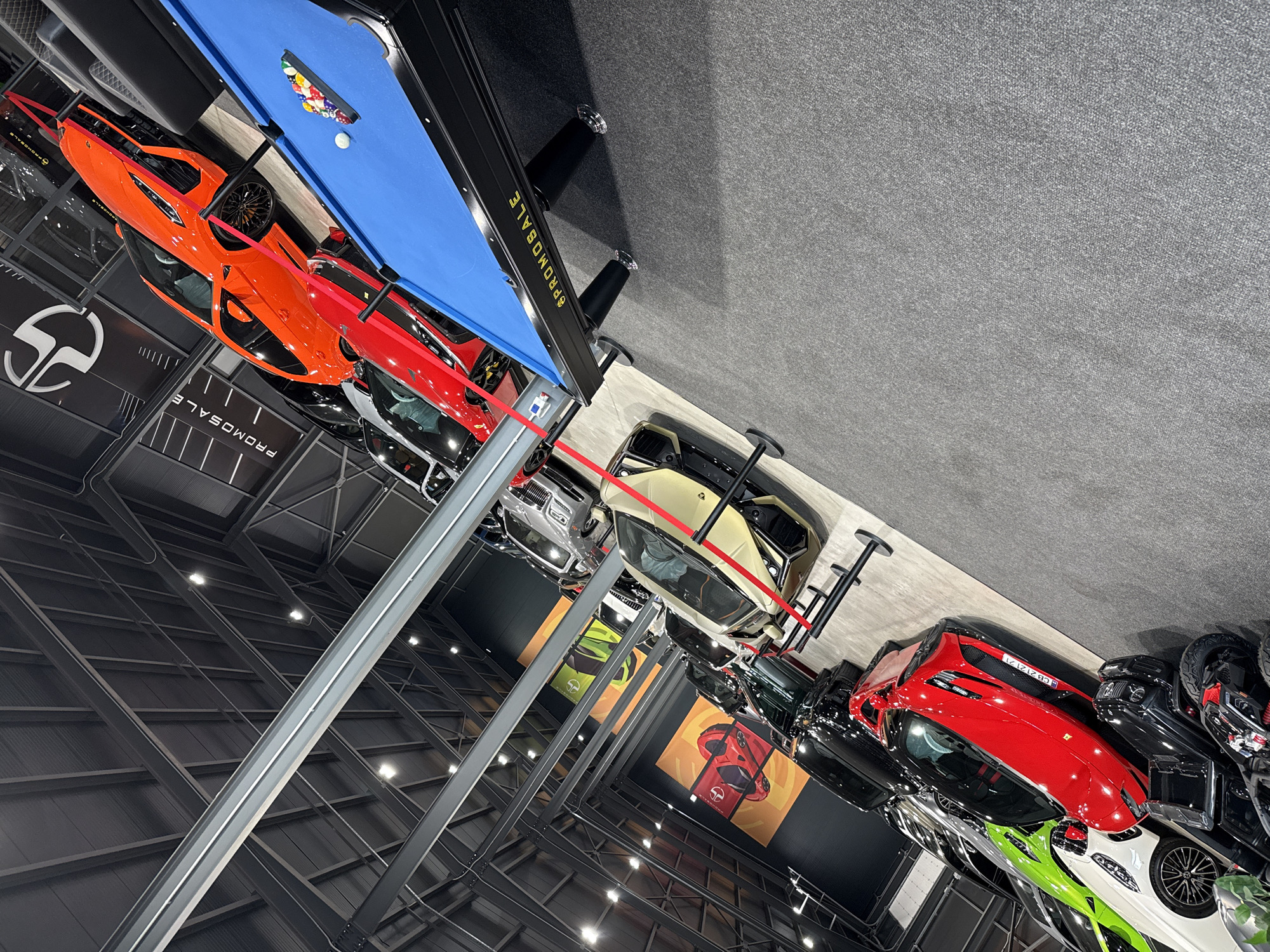 This image captures a wide-angle view of a luxury car showroom or exhibition hall, likely located in Plovdiv, Bulgaria, with a variety of high-end vehicles on display. The overall scene is brightly lit by artificial lights, illuminating the polished surfaces of the cars and the industrial-style ceiling structure with metal beams.
In the foreground, a blue-felt pool table is prominently featured, equipped with a rack of colored pool balls and a white cue ball. The side of the pool table bears the golden text "PROMOSALE." Below and around the pool table, numerous sports cars are showcased. An orange sports car and a red sports car are most visible, with parts of a white, lime green, and other dark-colored vehicles also discernible. A red car has a license plate "CB2121Z."
The setting features dark gray carpeted flooring near the pool table, transitioning to lighter, reflective floor surfaces where the cars are parked. Many cars are roped off with red barriers, indicating display areas. Large black walls feature white stylized "S" or "SP" logos and the text "PROMOSALE" on banners, emphasizing a sales event. Further into the background, other luxury vehicles, including a tan/gold SUV (possibly a Lamborghini Urus) and several black cars, are visible. Large posters with car illustrations in orange and green are mounted on the walls. The scene conveys a dynamic and upscale atmosphere associated with high-performance automotive sales. No people are visible in the frame, allowing the vehicles and showroom design to be the central focus.