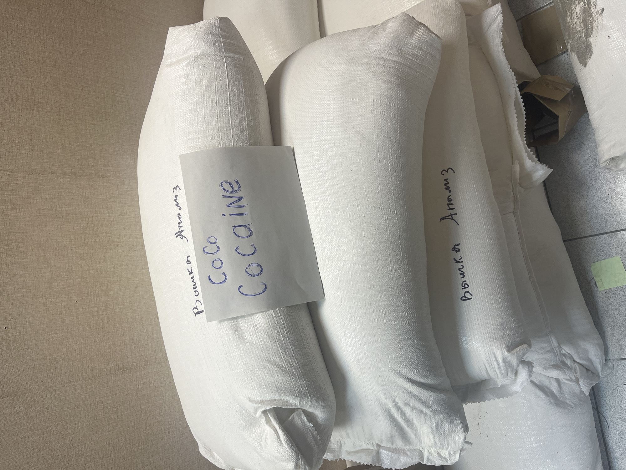 The image shows several white, woven bags stacked against a wall and on a tiled floor. One bag has a piece of paper taped to it with the word "Cocaine" written in blue ink. The word "Вышка Анализ" is written in Russian on at least two of the bags. Part of a cardboard box is visible near the bags. There are no people in the image. The environment is a simple indoor space. There is no information in the image to confirm its location in the city of Ulyanovsk, Russia.