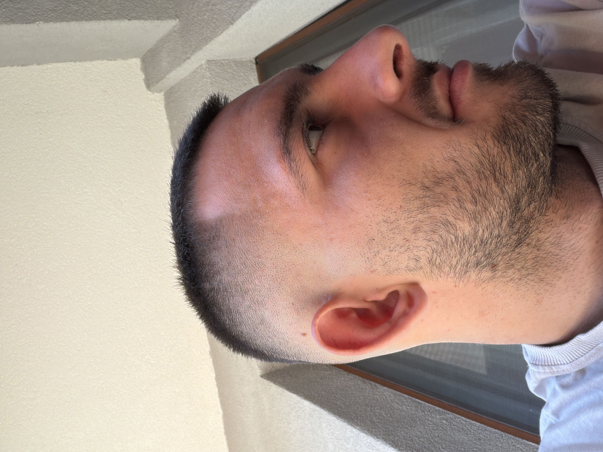 A close-up profile shot of a man's head. He has short, dark hair, a trimmed beard, and light skin. His nose, lips, and ear are visible. The background consists of a light-colored wall and a window frame. The location in Plovdiv, Bulgaria is not discernible from the visual information provided.