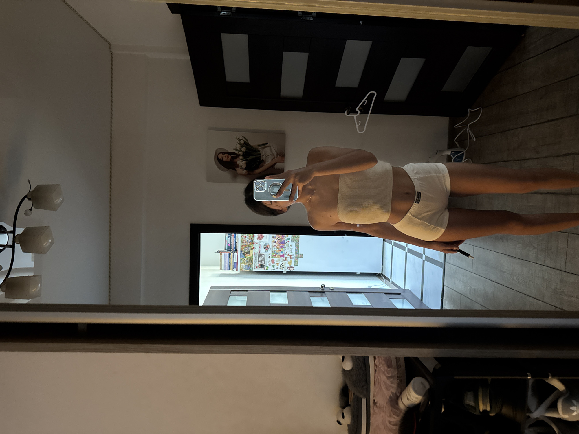 A young woman is standing in what appears to be a bedroom and taking a mirror selfie. She is wearing a white strapless crop top and white shorts. She is holding a phone in her right hand, and a black pen or stylus in her left. The room has wooden floors and white walls. In the background, reflected in the mirror, there is a doorway leading to another room, a black door, and a framed picture on the wall. There are also clothes and shoes visible in a closet area to the left. The lighting suggests it is indoors, possibly in the late afternoon or evening. The image does not provide any specific visual cues that would allow for identification of the location within Chișinău, Moldova.