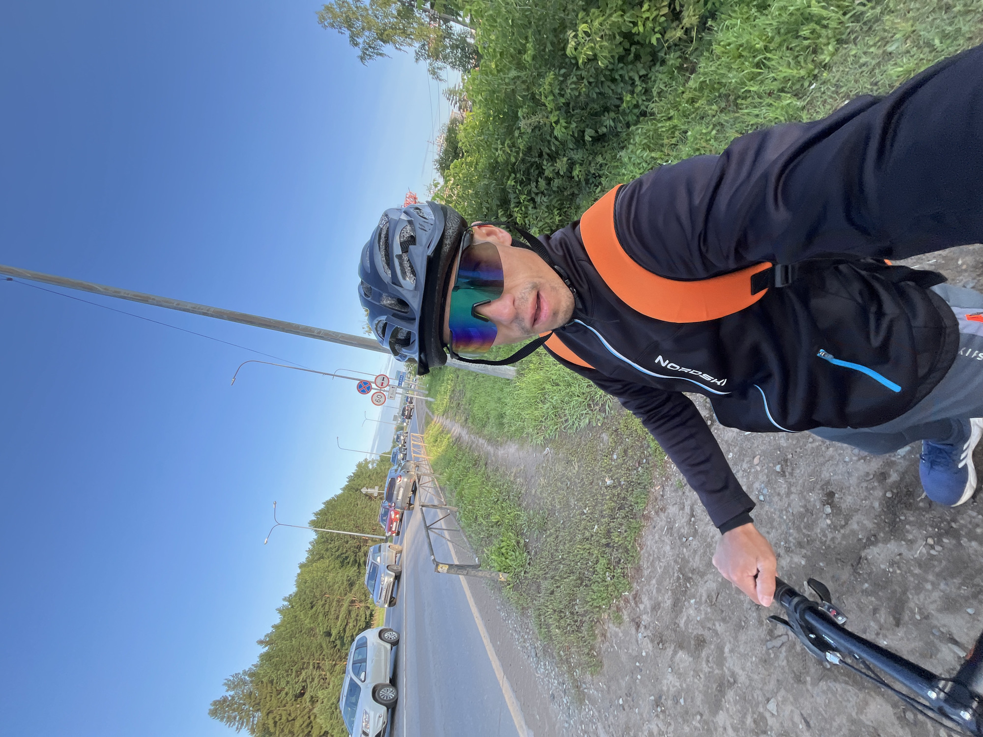 A man wearing a black cycling helmet and iridescent cycling glasses is taking a selfie while riding a bicycle. He is wearing a black long-sleeved cycling jacket with blue accents and an orange backpack. The bicycle handlebars are visible in the foreground. The man is on a dirt path next to a paved road with moderate traffic, including SUVs and cars. There are trees and green foliage on the right side of the road, and a dense line of trees in the background. The sky is clear and blue. Several streetlights and traffic signs are visible along the road. The scene appears to be on the outskirts of the city of Chaikovsky, Russia, based on visual cues of the road and vegetation typical of the region.