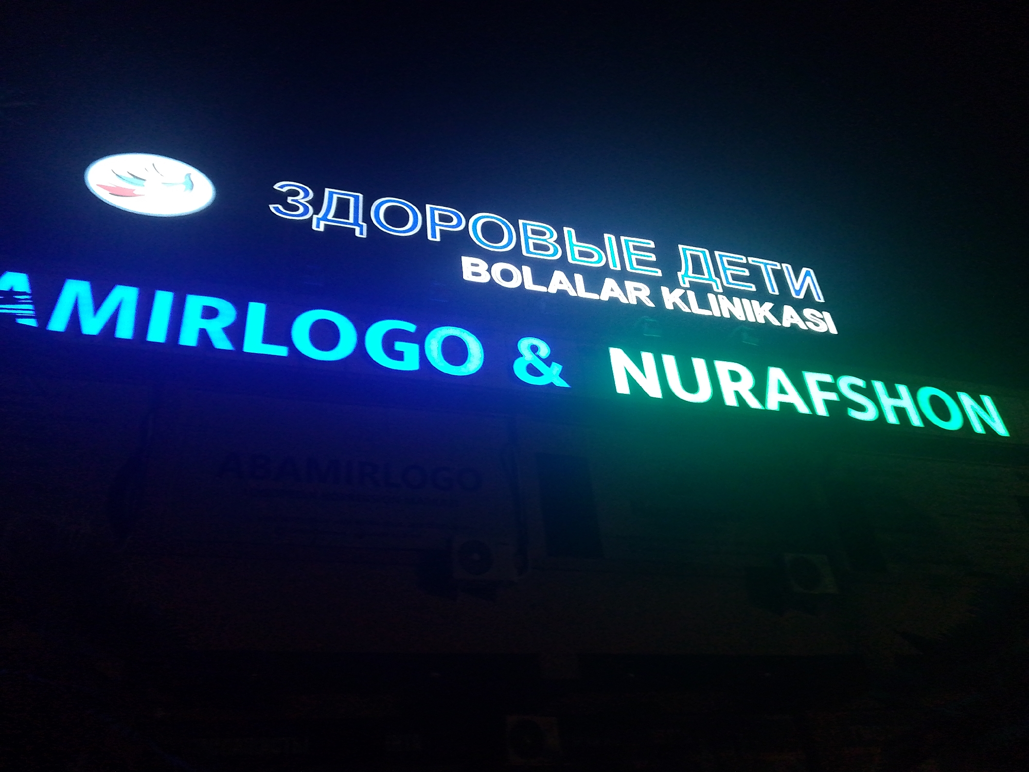 A nighttime exterior shot of a building facade in Bukhara, Uzbekistan, features illuminated signage. The top line of the sign displays "ЗДОРОВЫЕ ДЕТИ" in blue and white neon, translating to "Healthy Children." Below this, in smaller white lettering, is "BOLALAR KLINIKASI," meaning "Children's Clinic." A logo, resembling a smiling moon or cloud with closed eyes and pink blush marks, is positioned to the left of the top text. The bottom portion of the sign consists of large, bright green neon letters spelling out "& NURAFSHON," with a blue neon "AMIRLOGO" above it, though "AMIRLOGO" is less illuminated. The building itself is constructed of brick or stone and appears to be a multi-story structure. Visible on the facade are two air conditioning units, one in the middle and another towards the right side of the frame. The lighting is predominantly from the neon signs, casting a blue and green glow on the dark surroundings and the building's texture.