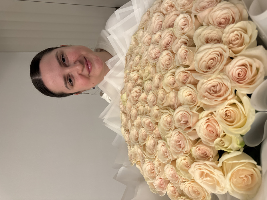 A woman is featured in a close-up, half-portrait style photograph, presenting or holding a remarkably large bouquet of roses. Her dark hair is neatly pulled back, revealing a serene face with a gentle, closed-mouth smile. She wears a white top, partially visible on the left side of the frame.
The dominant feature of the image is the enormous bouquet of roses, which fills the majority of the right and lower portions of the photograph. The roses are uniformly light in color, appearing as a blend of pale pink and creamy white hues, tightly packed together to create a dense, luxurious display. The bouquet is elaborately wrapped in multiple layers of crisp white and translucent paper, forming a ruffled, decorative collar around the extensive floral arrangement.
The setting appears to be indoors, against a plain, light gray wall that provides a simple and uncluttered background, allowing the subjects to stand out. The activity captured is likely a moment of celebration, appreciation, or the presentation of a significant gift. The woman's expression conveys a sense of quiet joy or contentment. There are no specific cues for the time of day or weather as it's an indoor scene. No visible text is present in the image. The location context provided for this media file is Dubai, United Arab Emirates.