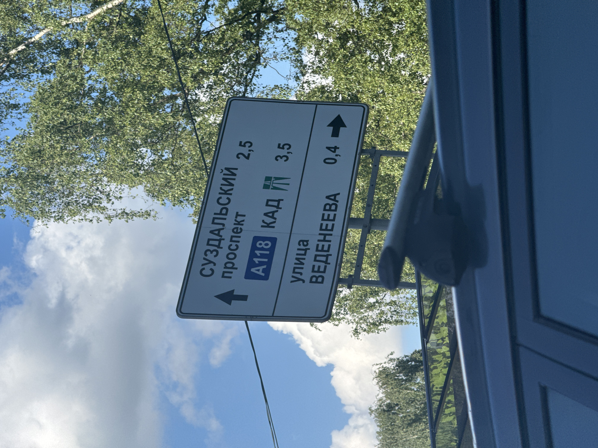 The image is oriented vertically, capturing a road sign against a backdrop of a bright sky and lush green trees. The sign is white with black text and symbols, framed by a dark border, indicating directions and distances to local roads and a major highway.
Visible text on the sign, from top to bottom, includes:
* "СУЗДАЛЬСКИЙ проспект" (Suzdalsky Prospekt) followed by "2,5" (kilometers) and a left-pointing arrow.
* A blue rectangle with "A118" inside, next to "КАД" (KAD, referring to the Saint Petersburg Ring Road), followed by "3,5" (kilometers) and a left-pointing arrow.
* "улица ВЕДЕНЕЕВА" (Ulitsa Vedeneeva) followed by "0,4" (kilometers) and an upward-pointing arrow (indicating straight ahead).
The background features a clear blue sky with scattered white clouds and the dense foliage of green trees, suggesting a sunny day. A glimpse of a grey railing or support structure is visible on the right side of the sign. To the far right and partially obscuring the view, there is a dark, possibly metallic, structure, which appears to be part of the vehicle from which the photo was taken, possibly a window frame or part of a roof rack/door.
The Cyrillic text and specific road names like "КАД" and "Suzdalsky Prospekt" strongly indicate the location is Saint Petersburg, Russia. The scene depicts a typical outdoor setting with road infrastructure, likely taken during daylight hours on a clear weather day.