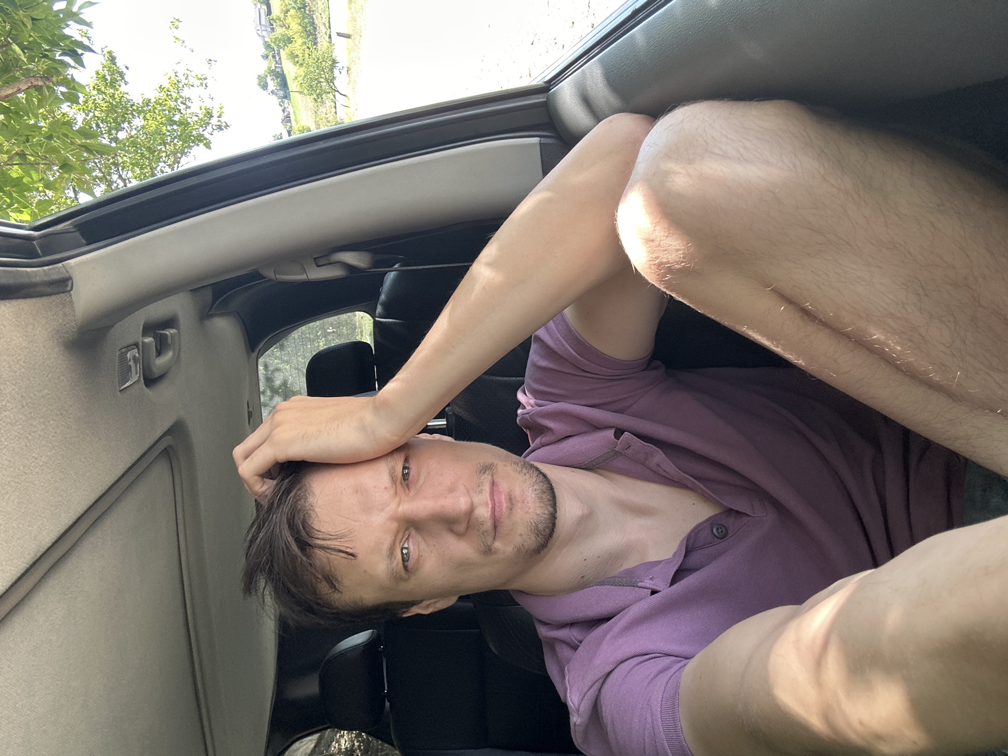 A man with short, dark hair and a short beard sits in the passenger seat of a vehicle, looking towards the camera. He is wearing a purple polo shirt and has his left hand on his forehead. His right arm is resting on his knee, which is visible in the foreground. Sunlight streams into the car, casting a shadow on his leg. The background outside the car shows a grassy hillside with scattered trees and houses. The scene appears to be outdoors, possibly in a rural area. The image was taken on a sunny day.