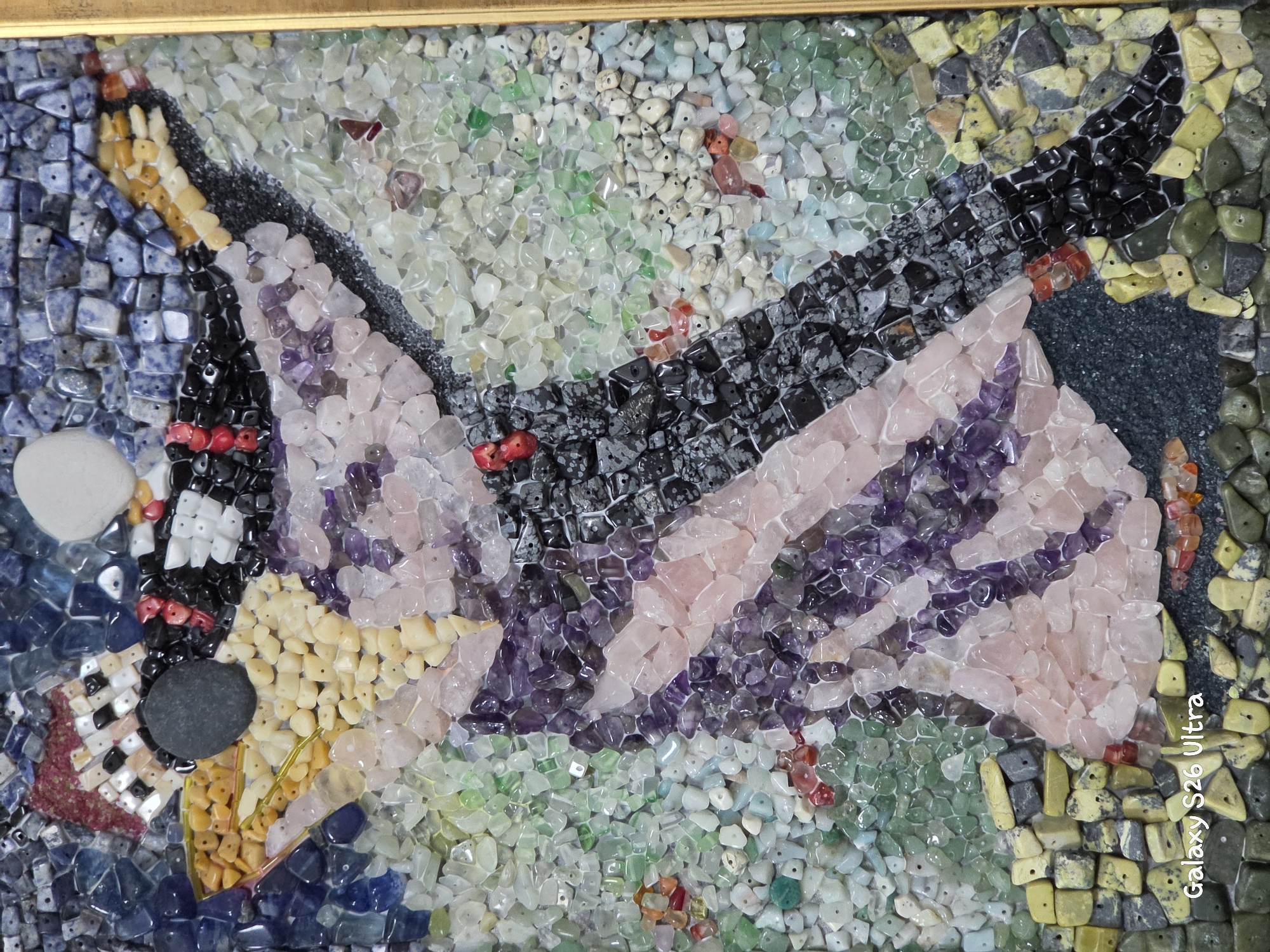 This is a close-up, top-down, vertical shot of a mosaic artwork. The mosaic is made of small, irregular pieces of polished stones and minerals in various colors, including shades of blue, purple, pink, green, yellow, black, and white. The artwork depicts a stylized figure in what appears to be a dancing pose, with flowing garments. The figure is predominantly rendered in shades of pink and purple for the clothing, with black and grey for the head and parts of the outfit. The background is a textured mosaic of blue, grey, and green stones. A small, round, white stone is positioned in the upper right quadrant, possibly representing the moon or a sun. The bottom left corner of the image displays a watermark: "Galaxy S26 Ultra". There is no discernible location in Plovdiv, Bulgaria, evident in the image; it is a studio shot of a standalone artwork.