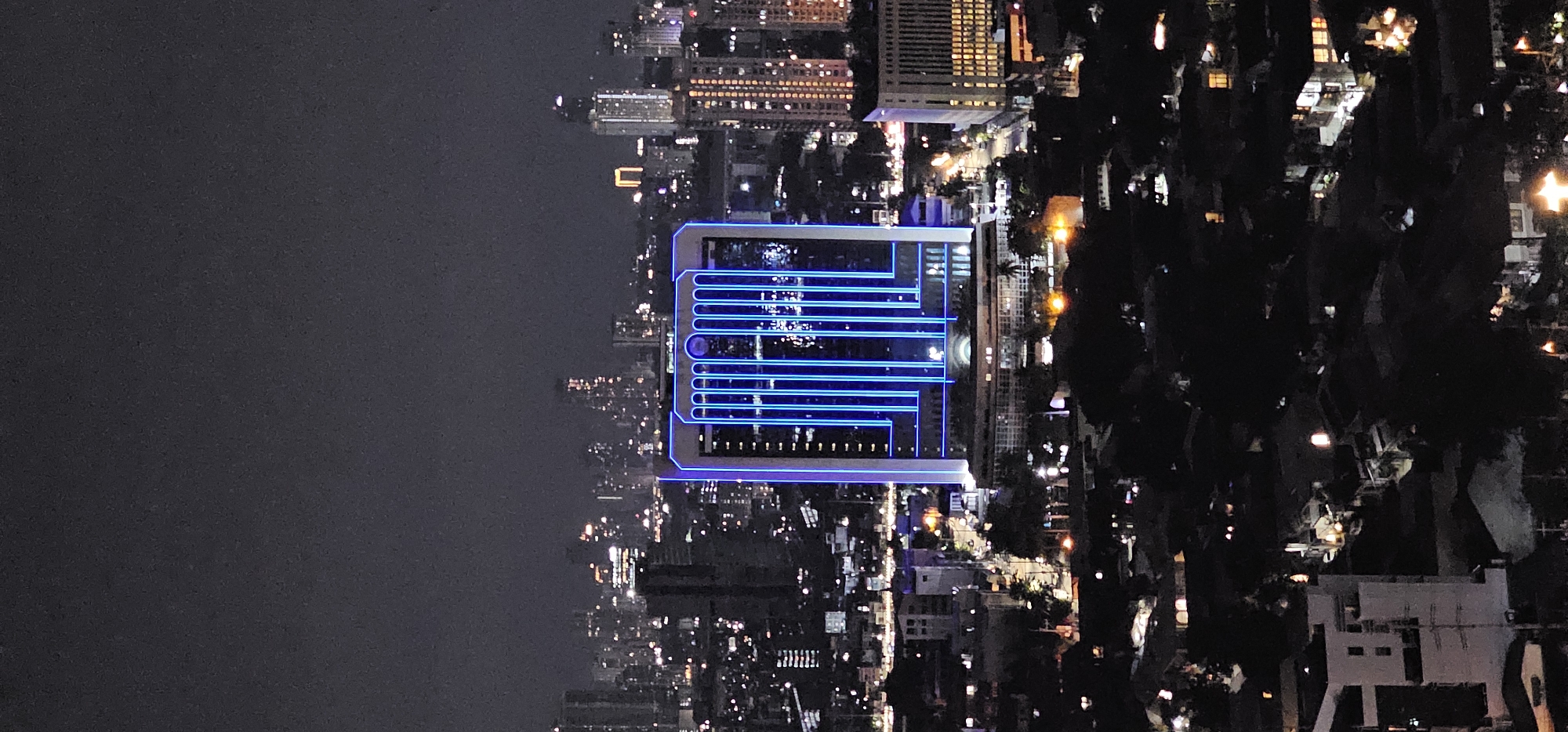 This image captures a nighttime aerial view of the Makati cityscape in the Philippines. The focal point is a modern skyscraper illuminated with striking blue vertical lines of light, standing out against the dark sky and the surrounding urban landscape. Numerous other buildings, varying in height and illuminated by a multitude of lights, form a dense urban sprawl. The foreground reveals a lower-lying residential area with darker structures and scattered lights, suggesting a contrast between commercial and residential zones within the city. The overall impression is one of a vibrant and active metropolitan area at night.