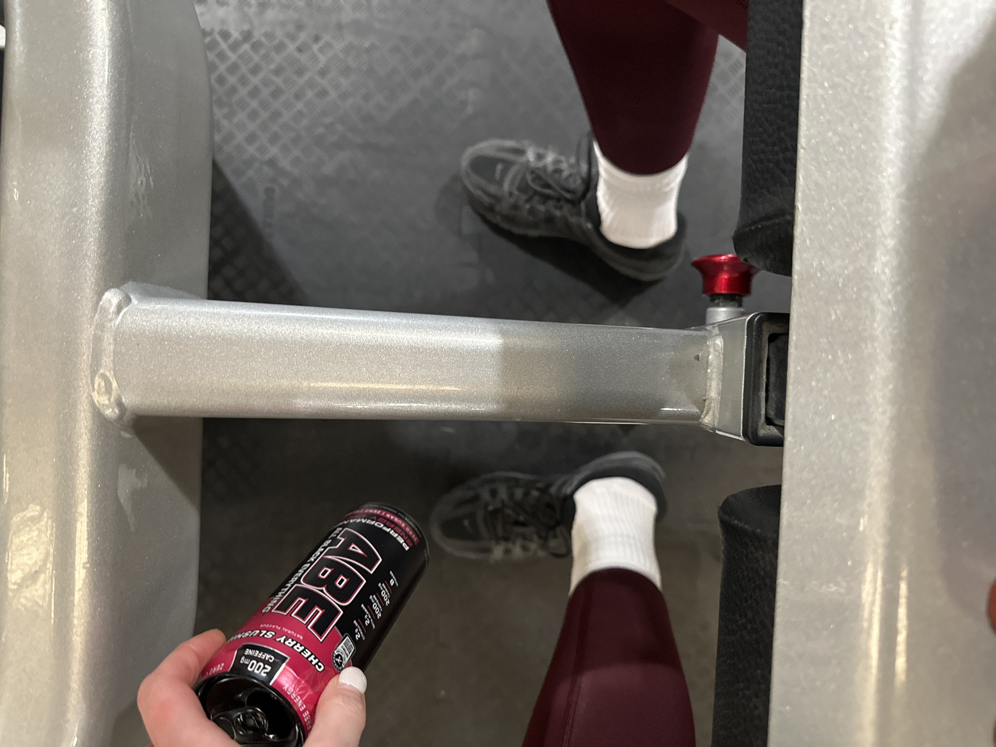 The image shows a person at a gym. The person is wearing maroon leggings, white socks, and black sneakers. They are holding a black and red can of "ABE All Black Everything" energy drink. The can states it has 200mg of caffeine and is cherry slush flavored. The person appears to be sitting on a piece of gym equipment. The flooring of the gym is gray with a checkered pattern. The gym equipment is gray metal. The location is a gym, but the city of Tunbridge Wells, United Kingdom, cannot be determined from the image.