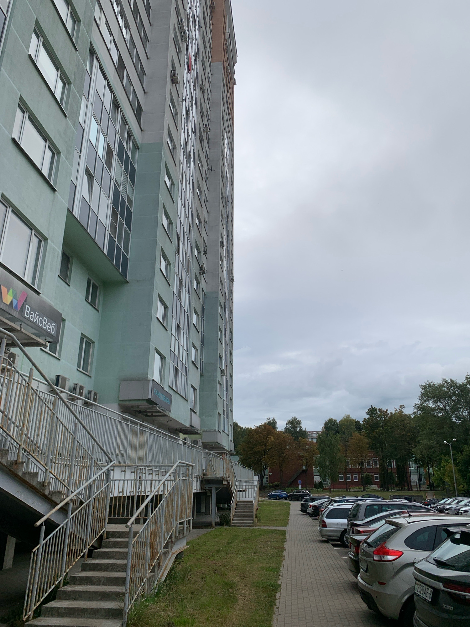 An exterior, eye-level, medium shot captures the side of a tall, light green apartment building in Minsk, Belarus. The building's facade features numerous windows, some with reflective glass, and several air conditioning units are mounted externally. To the left, a metal staircase with a handrail leads up to an entrance. Above the staircase, a sign displays the text "ВайсВеб" with a colorful logo. Further down, another sign reads "КАНЦТОВАРЫ". Adjacent to the building, a paved walkway runs alongside a parking lot filled with various cars. Trees with some autumn foliage are visible in the background, and the sky is overcast.