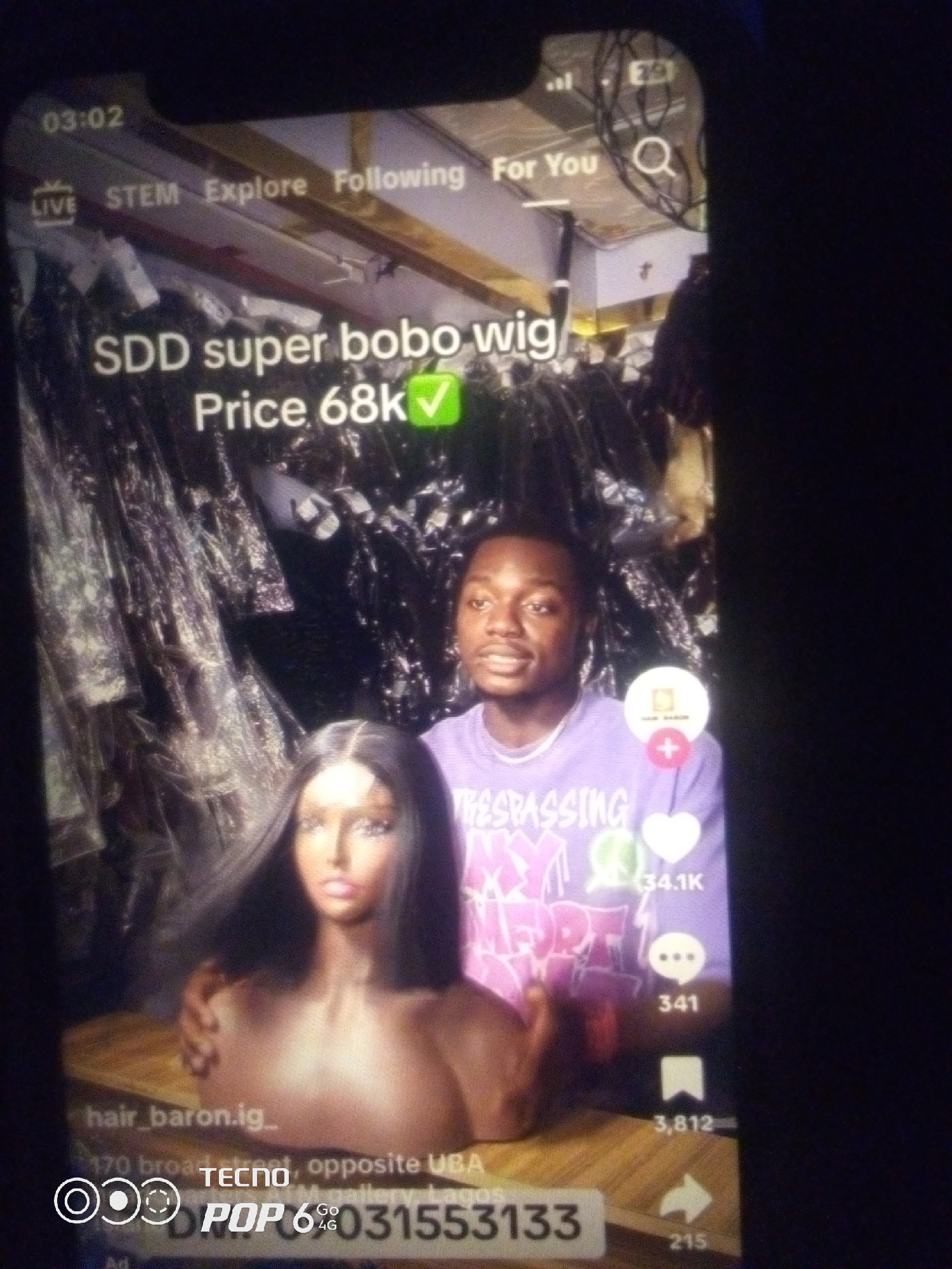 The image captures a close-up, vertical shot of a smartphone screen displaying a social media video. The video features a mannequin head with dark hair in the foreground, positioned to the left. To the right of the mannequin is a young man wearing a purple t-shirt with text on it. The background appears to be a retail space with racks of clothing or merchandise. Overlaying the video content are user interface elements common to social media platforms, including a timestamp "03:02" at the top left, navigation tabs like "LIVE," "STEM," "Explore," "Following," and "For You" at the top. Text on the video reads "SDD super bobo wig Price 68k" with a green checkmark. Below the video, social media engagement metrics are visible, including icons for likes, comments, and shares, with accompanying numbers like "34.1K" and "341". At the bottom of the screen, an advertisement for "TECNO POP 6 4G" is displayed, along with contact information. The overall image quality is low, with motion blur and reflections indicating it was taken in low light conditions, possibly at night. The text "hair_baron.ig_" and an address "170 broad street, opposite UBA" suggest the location is within Lagos, Nigeria, not Howayi.