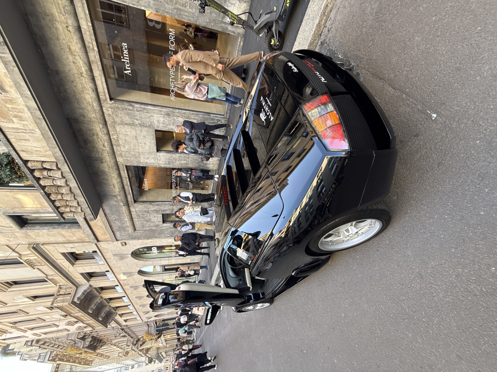 An urban street scene in Milan, Italy, captured during daylight, shows a black sports car parked on the right side of the road with its passenger-side scissor door open. The car, which appears to be a Lamborghini Murciélago, features distinct taillights, side vents, and silver multi-spoke wheels. Text visible on the car's rear includes "AUTODIAVOLO" and "ALCHYMIA". The interior, visible through the open door, has light-colored seating.
To the left of the car, a wide sidewalk is lined with multi-story buildings featuring classic European architecture and large display windows. Several people are walking or standing on the sidewalk and inside the storefronts. One man in a light brown suit walks past the rear of the car. An electric scooter, marked with "GFEITE", is parked on the sidewalk near a building entrance.
Storefront signs and window displays reveal text such as "Arelinea" and "ARCHETYPE -- FORM" on one window, and "LAGO" on another. The setting suggests a bustling commercial area. The overall impression is one of everyday city life, with a luxury car drawing attention.