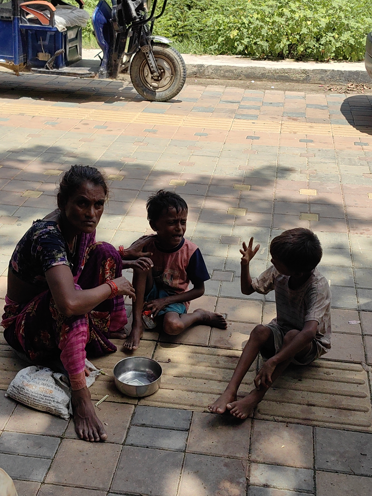 In Lumbini Bazaar, Nepal, three individuals, an adult female and two young children, are seated on a patterned paved surface. The adult female, wearing a dark floral print top and a pink and floral patterned skirt, is positioned on the left. She has bangles on her left wrist and appears to be looking towards the viewer. To her right, a younger child, wearing a light-colored t-shirt and dark shorts, is crying. The third individual, a slightly older child in a striped t-shirt and shorts, sits to the right of the crying child. This child is gesturing with their left hand, with fingers extended. In front of the adult, a metal bowl contains some money, and a white cloth bag lies beside it. In the background, an electric rickshaw is visible, and beyond that, lush green vegetation lines the edge of the scene. The ground is paved with square tiles in varying shades of brown, beige, and gray, with tactile paving strips running horizontally across the image.