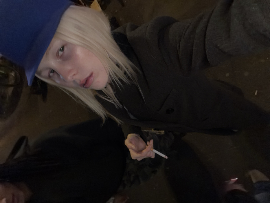 The image depicts two individuals in a dimly lit, likely outdoor, urban environment. In the foreground, positioned right of center, a person with pale skin and shoulder-length platinum blonde hair wears a royal blue, flat-brimmed hat and a dark grey, double-breasted coat. This individual holds a lit cigarette between the thumb and index finger of their right hand, with a visible wisp of smoke emanating from it. Their gaze is directed forward, towards the camera, with a neutral expression and slightly parted lips. Partially visible in the upper left background is a second person with dark hair, wearing a dark hooded sweatshirt and what appears to be camouflage-patterned trousers. The ground is dark, possibly asphalt or concrete, with some minor reflections and small specks of debris. Indistinct objects, possibly including parts of a bicycle, are faintly discernible in the dark background. The specific location within the city of Paris, France, cannot be determined from the visual evidence presented.