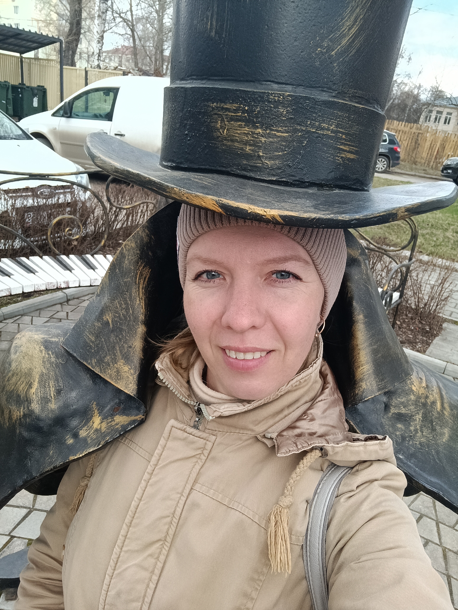 A woman with fair skin, light blue eyes, and a cheerful smile poses for an outdoor photo, playfully aligning her head beneath a large, decorative top hat sculpture. She wears a beige knitted beanie, a warm beige quilted jacket with a zipped collar, and a light-colored strap from a shoulder bag. A small gold hoop earring is visible in her right ear.
The prominent sculpture is an oversized, antique-style top hat, black with distressed gold accents, suggesting an aged or theatrical prop. It rests on a dark, stylized structure that appears to be part of a larger figure or cloak. In the background, to the left, a segment of a large outdoor piano keyboard sculpture with white and black keys is visible, along with an ornate metal fence featuring spiral designs.
The outdoor setting includes bare trees, indicative of late autumn, winter, or early spring. Several vehicles, including a white van and a dark car, are parked behind a light-colored wooden fence. Green dumpsters and distant buildings are also part of the backdrop under an overcast sky. The ground is paved with a gray, cobblestone-like pattern. The woman's warm attire and the cool-weather foliage suggest a chilly day. The overall scene conveys a pleasant, lighthearted moment in a public space, likely a park or plaza in Chaikovsky, Russia, possibly themed around music or art.