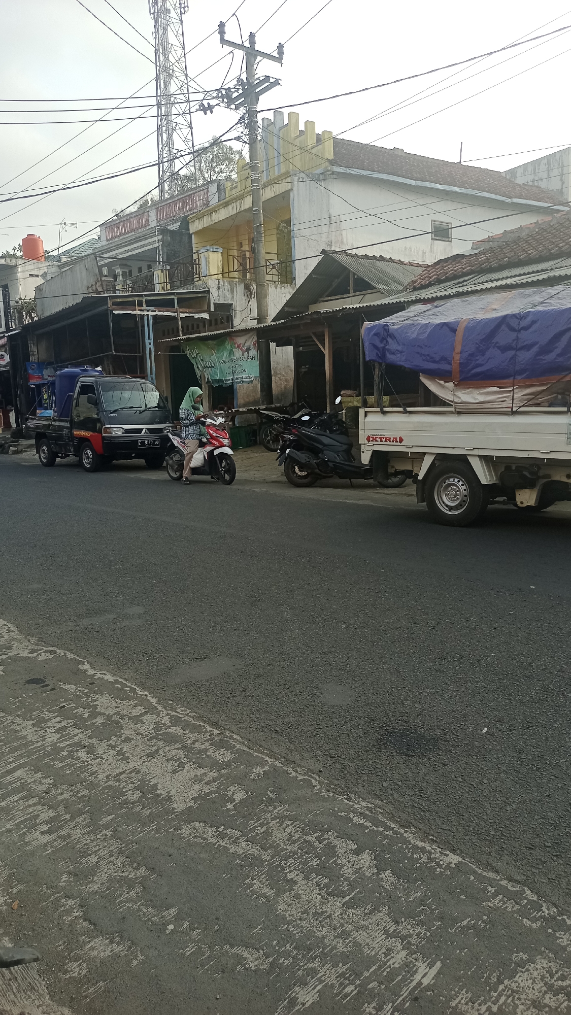 The image depicts a street scene in Karyamandala, Indonesia, likely a commercial area, with a focus on vehicles and buildings. In the foreground, the asphalt road shows signs of wear and tear. To the left, a black pickup truck is parked, with a person wearing a light green headscarf and checkered shirt riding a red and white motorcycle. The motorcycle is positioned next to the truck. To the right of the motorcycle, a black scooter is parked. Further to the right, a white flatbed truck is visible, its cargo covered with a blue tarp. Behind the vehicles, a row of buildings lines the street. These buildings appear to be a mix of shops and residences, with some exhibiting yellow and white facades and tiled roofs. A prominent telecommunications tower stands tall in the background, along with overhead electrical wires crisscrossing the sky. The sky is overcast.