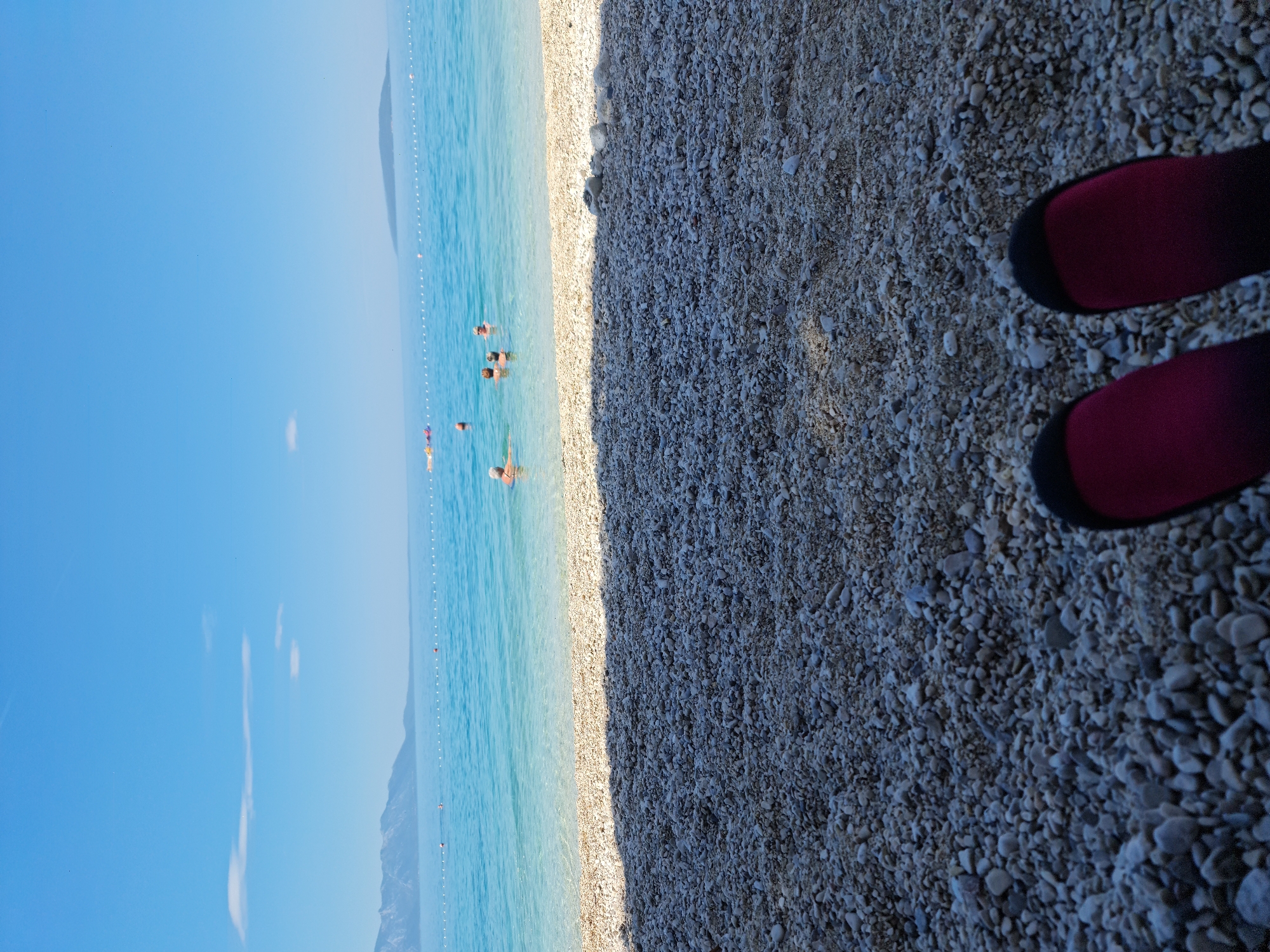 The image depicts a sunny day at a pebble beach in Općina Gradac, Croatia. The foreground is filled with grey and white pebbles, with the tips of two pink and black water socks visible in the bottom right corner, suggesting someone is sitting on the beach. The midground shows the clear, turquoise water of the Adriatic Sea, with a white rope barrier marking a swimming area. Several people are visible in the water, some swimming and others relaxing. In the background, a mountain range stretches along the coastline under a bright blue sky with a few wispy clouds. A small, distant island is also visible on the horizon.