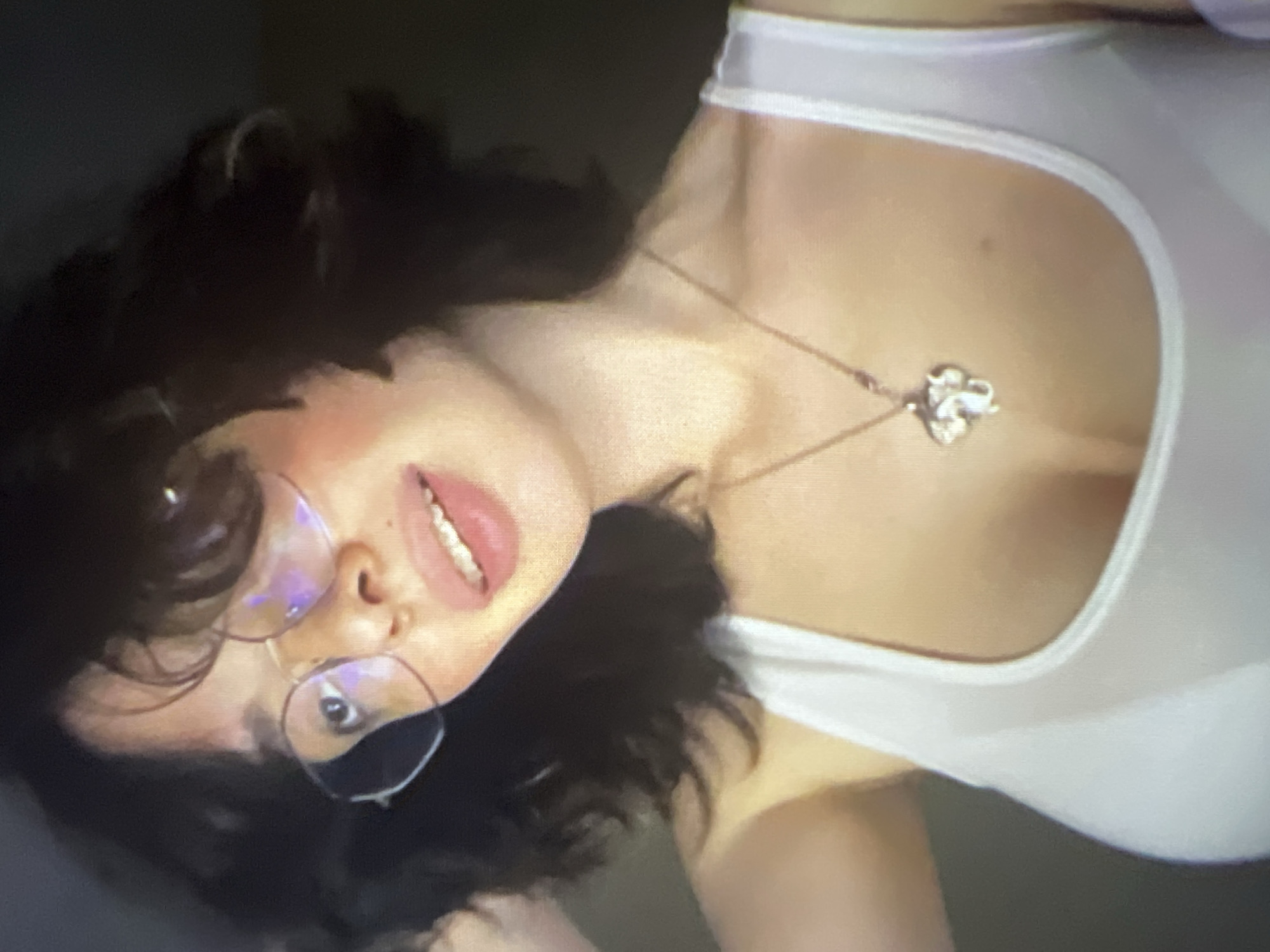 A close-up, low-angle shot shows a young woman wearing glasses and a white tank top. She has dark, wavy hair and is looking at the camera with her mouth slightly open, revealing her teeth. A silver pendant, shaped like an elephant, hangs from a necklace around her neck. The background is dark and out of focus. There is no information about the location in Pyatigorsk, Russia.
