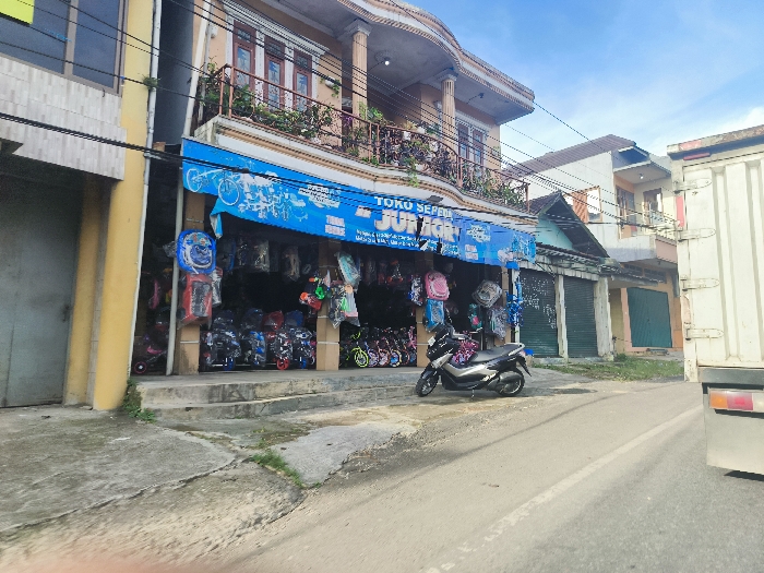 The image shows a street scene in Cikalong, Indonesia (latitude: -6.739, longitude: 107.439). The street is paved and has a white dividing line in the center. A black motorcycle is parked on the right side of the street. In the background, a white truck is partially visible. On the left side of the street is a store with a sign that says "TOKO SEPEDA JAYA," which means "Bicycle Shop JAYA" in Indonesian. The store appears to sell bicycles and other related items. There are several other buildings in the background, including a small residential building with a green roof. The sky is clear and blue. The buildings are all painted in light colors, and the area appears to be in a residential neighborhood. There is a small amount of green vegetation visible in the background. There are no people in the image.