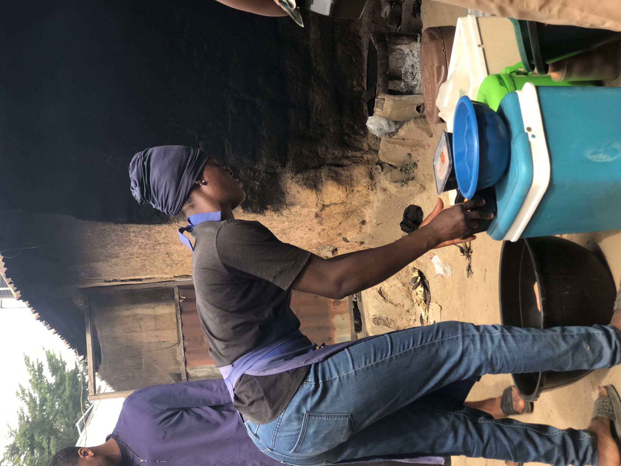 This image captures a daytime scene, likely in an outdoor or semi-outdoor setting, possibly a domestic compound or small-scale food preparation area in Jos, Nigeria. The ground is composed of sand or dirt.
The central subject is a person with dark skin, dressed in a dark grey t-shirt, blue denim jeans, and a blue headwrap. A blue piece of fabric is tied around their waist, resembling an apron or sash. They are positioned in a leaning posture, looking upwards and to their right, with their left arm extended. Their hand appears to be interacting with items placed on a light blue plastic cooler or storage box that has a white lid. On top of this cooler, a bright blue plastic bowl and a small, square, dark-colored object (possibly a scale or a container) are visible. This arrangement suggests the individual is engaged in weighing, measuring, or organizing goods, perhaps food items.
In the immediate vicinity of the central subject, a large black basin or bucket rests on the ground near their feet. Partially visible in the bottom left, another person wears a purple long-sleeved top and a beaded necklace. To the far right, the lower legs and feet of a third person, wearing dark sandals, can be seen.
The background shows a rustic environment with rough, dark walls, possibly stained by smoke, indicating a cooking or hearth area with visible ash or charcoal. Further back, portions of a corrugated metal structure and green foliage (trees) are discernable under what appears to be an overcast or shaded sky. The overall scene depicts a mundane, everyday activity within a simple, functional setting. There is no visible text in the image.