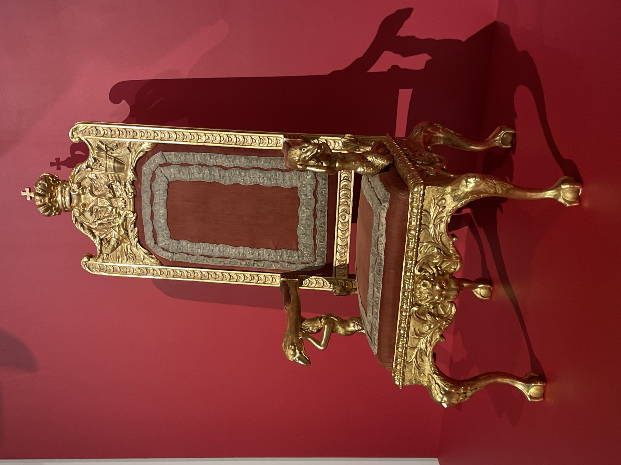 A gilded throne-like object is displayed on a red wall. The object is adorned with intricate carvings and embellishments and appears to be an antique. It is likely located in a museum or historical setting in Saint Petersburg, Russia, given the style and opulence of the object.
