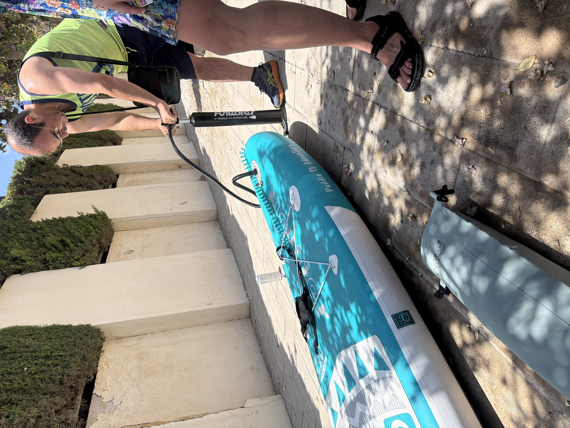 The image shows a man pumping up a stand-up paddleboard (SUP) outdoors. The man, wearing a neon green tank top and navy shorts, is using a black hand pump connected to the valve of the partially inflated blue and white SUP. The SUP is lying on a paved surface, likely a patio or walkway. In the background, there are cream-colored walls and a row of manicured green bushes. The sunlight casts shadows on the ground. A second, deflated paddleboard is visible in the foreground, on the left. A person's legs and feet are visible on the right side of the image, one wearing black sandals. The text "Paddle to Adventure" is visible on the SUP. The scene appears to be in Alicante, Spain, suggested by the style of architecture and vegetation.