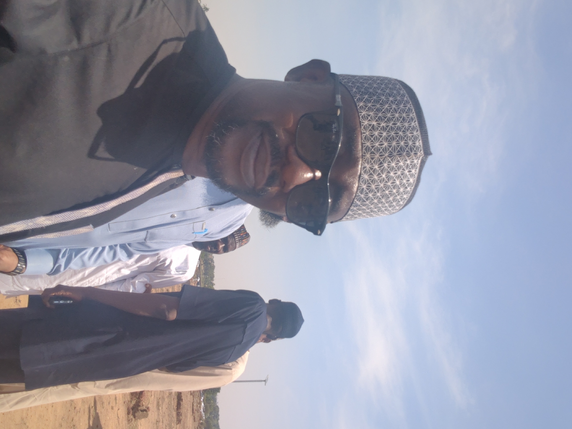 The image captures a group of men outdoors in Sardauna, Nigeria. One man is in the foreground, wearing a patterned kufi, sunglasses, a dark top, and a light blue shirt. Two other men are partially visible behind him, one in a white shirt and another in a dark blue top. The background shows a clear sky and dry, grassy land, suggesting a field or open area. There is a metal pole in the distance and a few other people at an unidentifiable distance. One man is holding a mobile phone in his hand.