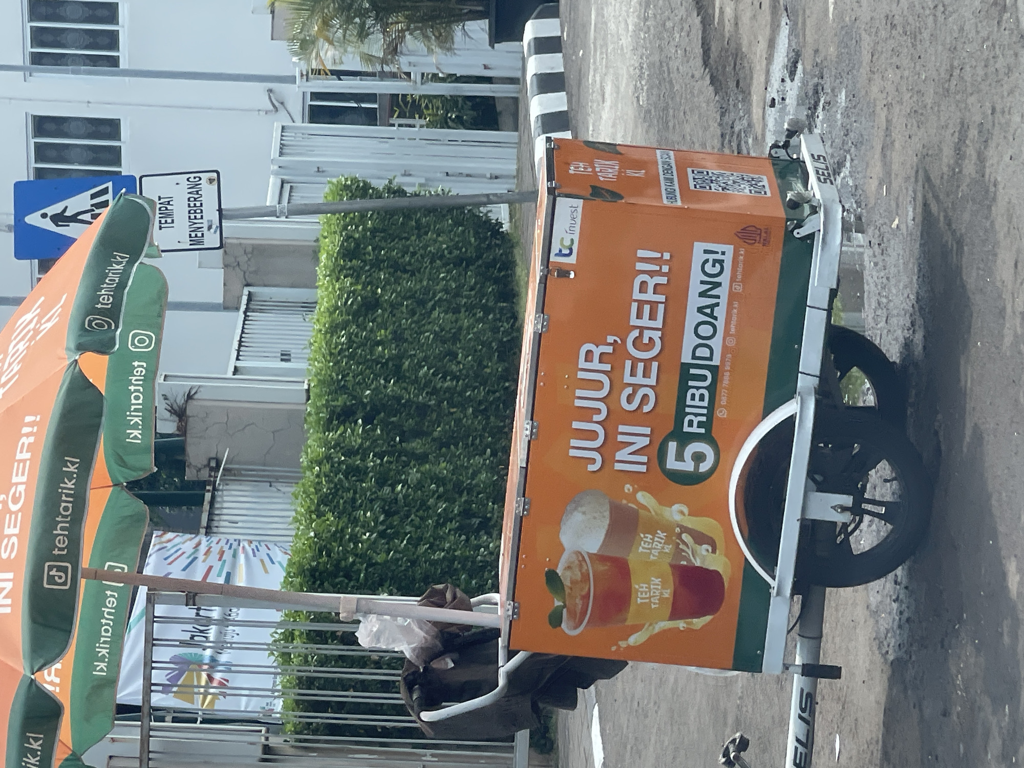 This is a medium shot, outdoor, eye-level, still photograph of a mobile beverage cart selling "Teh Tarik" in South Jakarta, Indonesia. The cart, which is orange and green, is parked on a paved surface, possibly a sidewalk or street. It is equipped with a large umbrella, also orange and green, providing shade.
Prominently displayed on the side of the cart are advertisements for "Teh Tarik KL" (Kuala Lumpur Teh Tarik). The text on the cart reads "JUJUR, INI SEGER!!" which translates to "Honest, this is refreshing!!" and "5 RIBU DOANG!" meaning "Only 5 thousand!". There are also images of two cups of "Teh Tarik" on the cart, one appearing to be a traditional milk tea and the other a lighter colored drink. A QR code and contact information are also visible.
To the left of the cart, a black backpack or bag is hanging on the handle. In the background, there is a dense green hedge and a white building with windows. A pedestrian crossing sign is visible above the umbrella, indicating a designated crossing area. The overall scene suggests a typical street vendor setup in an urban environment.