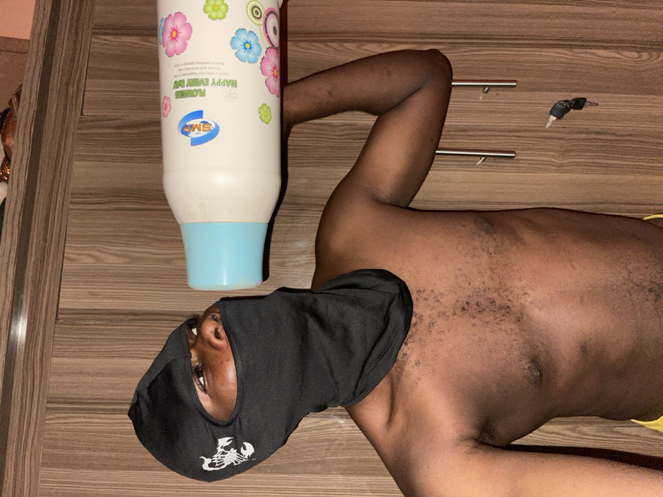 An individual, appearing male and dark-skinned, is positioned indoors within the city of Muchiya, Nigeria. The subject wears a black balaclava featuring a white scorpion logo on the left side, exposing only the eyes, nose, and mouth. The upper torso is bare, displaying a chest with dark hair. A yellow garment is partially visible at the bottom edge of the frame. The individual holds a large, white, opaque plastic bottle with a light blue cap, positioning the cap against their mouth as if consuming its contents. The bottle's label displays colorful floral graphics alongside the text "SMP," "FLOWERS," and "HAPPY EVERY DAY." The background consists of a vertical wooden paneling, against which a set of keys with a black fob hangs from a silver hook on the right side.