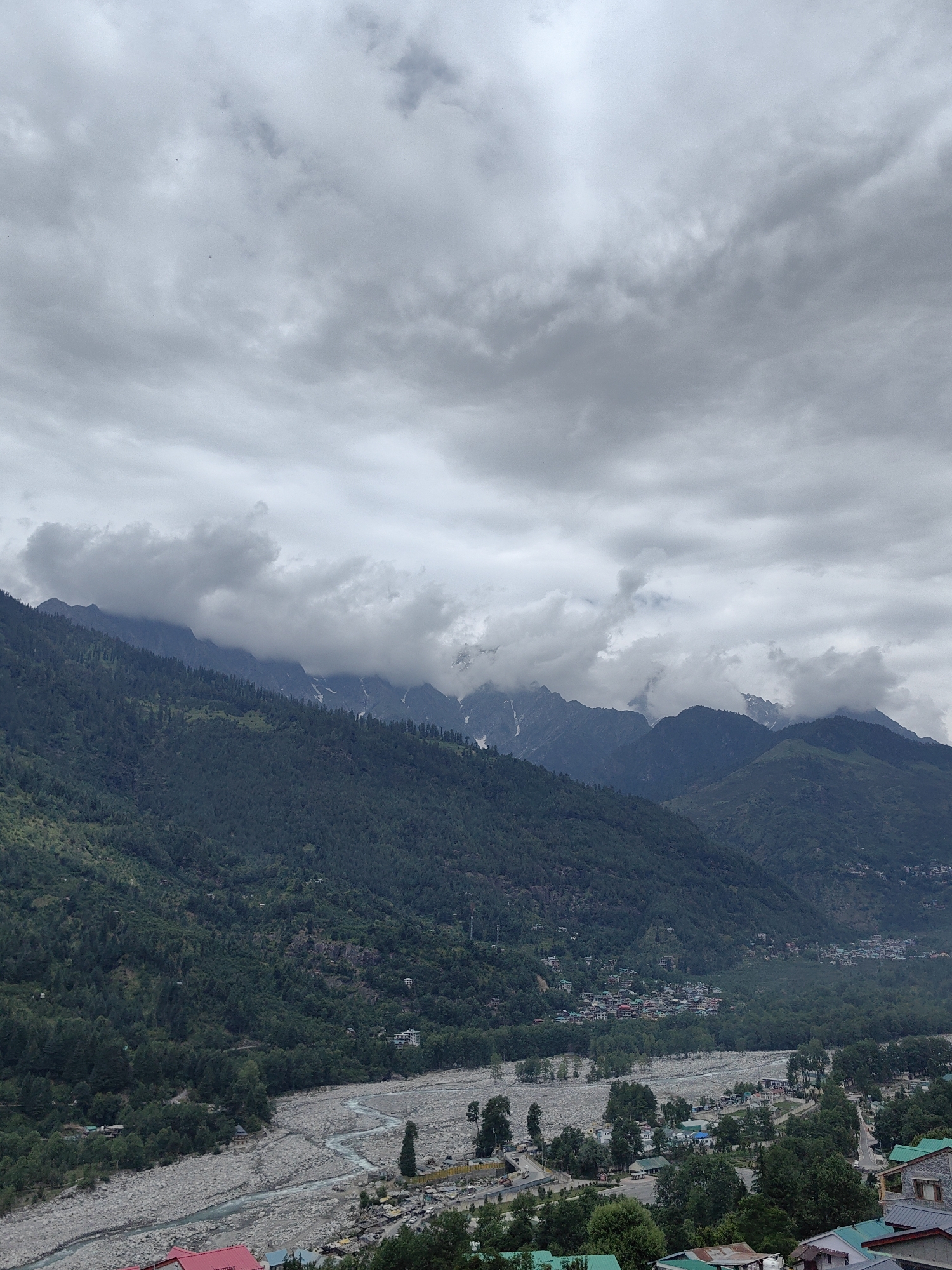 The image shows a wide landscape view of the city of Vashisht, India, nestled in a valley between lush green mountains. The sky is overcast with heavy grey clouds, hinting at impending rain or fog. In the foreground, a wide, rocky riverbed with a meandering stream cuts through the valley floor. Along the river and extending up the slopes of the mountains are dense patches of dark green trees, interspersed with scattered human settlements. Buildings with colorful roofs, typical of Indian architecture, are visible within these settlements, appearing as clusters of varying sizes. The mountains themselves are steep and covered in dense vegetation, with some higher peaks showing patches of snow or bare rock. The overall atmosphere is one of a natural, somewhat rugged, and populated mountain environment under a cloudy sky.