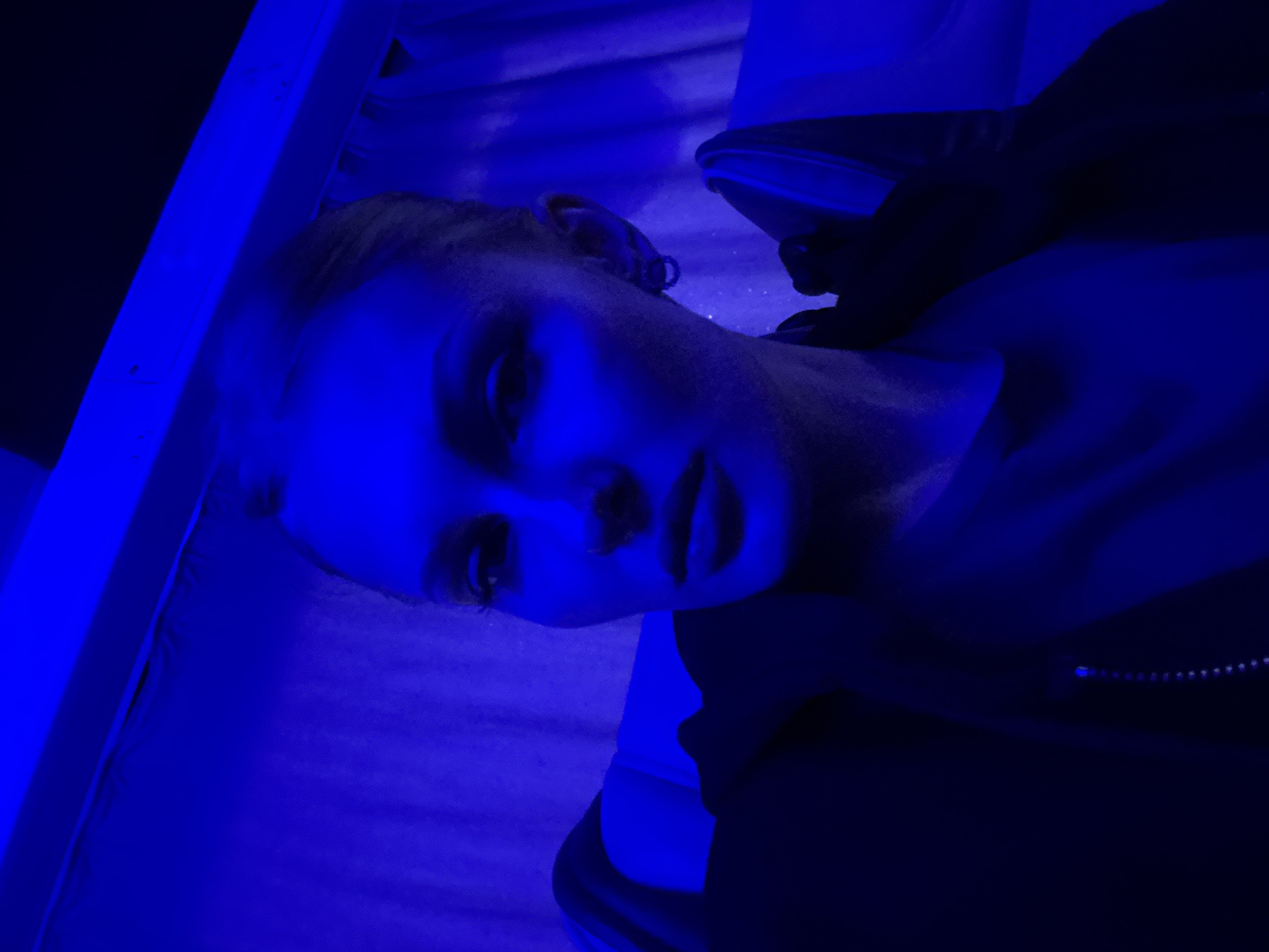 A close-up, low-angle, selfie-style photograph features a young woman with blonde hair pulled back, wearing dark clothing. She is illuminated by vibrant blue light, creating a dramatic and moody atmosphere. Her face is turned slightly towards the camera, with her gaze directed forward. The background consists of rippled fabric curtains, also bathed in the blue light, and hints of upholstered seating suggest an indoor environment, possibly a lounge or a vehicle. The overall impression is one of artificial lighting and a deliberate aesthetic choice. The location of Kazan, Russia, is not discernible from the visual information presented in the image.