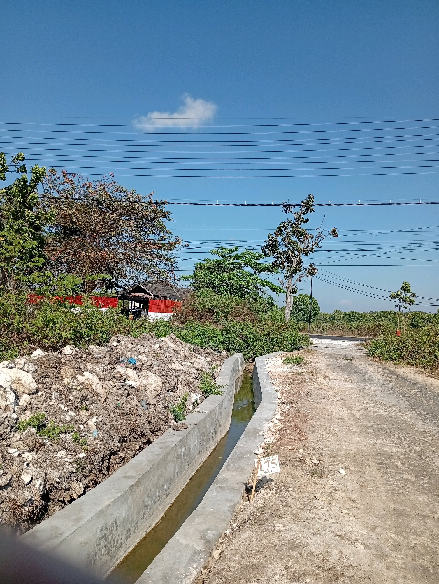 A view of a rural road in Socah, Indonesia, is captured under a clear blue sky. The road itself is paved and appears to be in a somewhat dilapidated condition, with visible cracks and loose gravel. To the left of the road, a newly constructed concrete drainage channel runs alongside it, filled with murky water. A pile of rocks and debris is situated between the drainage channel and a red and white structure, which appears to be a small building with a brown roof. Several trees with green and reddish-brown foliage are scattered throughout the scene, some taller and more established than others. Overhead, a network of electrical wires stretches across the sky, supported by poles that are not fully visible. In the foreground, a white sign with the number "175" and the word "METER" is staked into the ground next to the drainage channel. The overall impression is one of an undeveloped or semi-developed area, with ongoing construction or road maintenance activities.