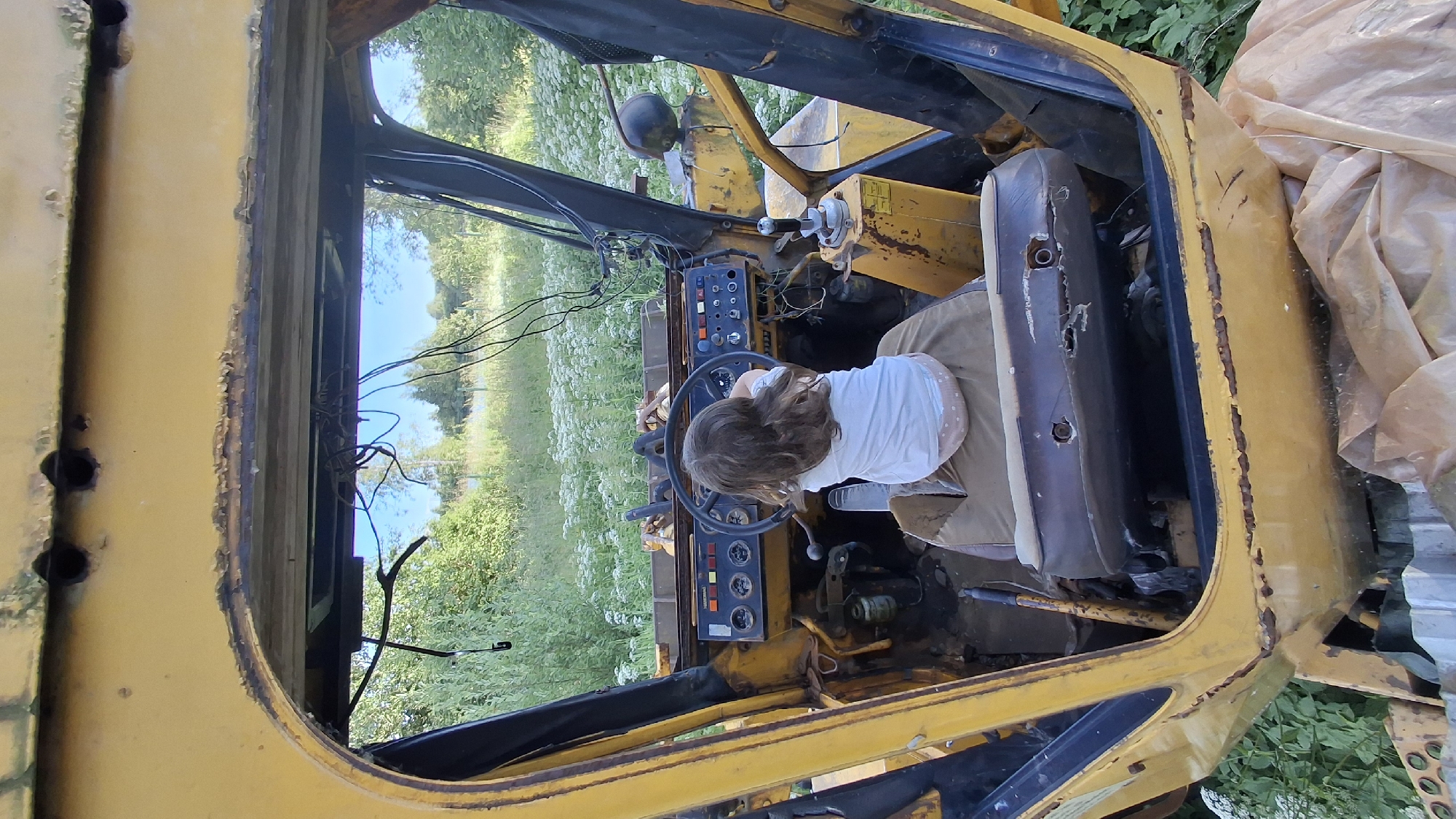 An overhead, slightly tilted view captures the interior of a dilapidated yellow construction vehicle, likely a tractor or similar heavy machinery, parked outdoors amidst a lush, green field. The vehicle's cabin is open to the elements, showing signs of age and disuse, including rust and exposed wiring. A young child, seen from behind, is seated in the driver's seat, gripping the steering wheel. The child is wearing a white t-shirt and light-colored bottoms. The surrounding environment suggests a rural setting, with abundant green foliage and white wildflowers stretching into the background under a bright sky, indicating daytime and fair weather. The scene evokes a sense of childlike curiosity and playfulness, with the child seemingly imagining operating the heavy machinery in this abandoned state. The location is identified as Innolovo, Russia. No visible text is present in the image.