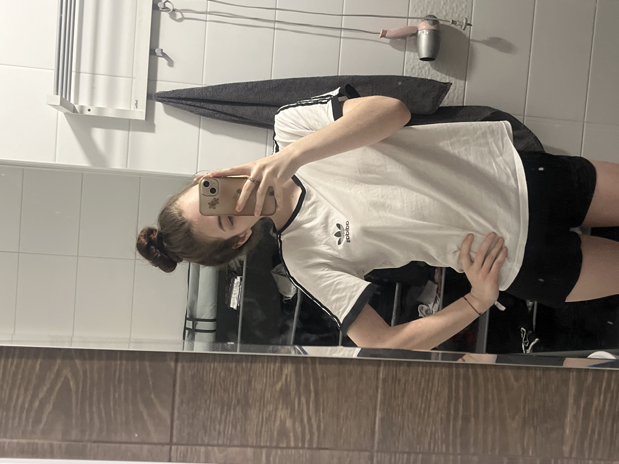 A female individual is captured in a mirror selfie, standing from mid-thigh to the top of her head. Her light-brown hair is gathered in a high, messy bun. She holds a gold-colored smartphone, consistent with an iPhone Pro model, in her left hand, partially obscuring her face from the nose upwards. A small floral decal is visible on the phone case, and a gold-colored ring is worn on her left ring finger. Her right hand rests on her hip. She is dressed in a white t-shirt featuring a black ringer collar, three black stripes on the sleeves, and an Adidas Trefoil logo on the left chest. Black shorts and a thin red string bracelet on her right wrist are also visible.
The environment reflected in the mirror displays a white tiled wall with horizontal grout lines on the right side. A white wall-mounted heating element or towel warmer is affixed to this wall, beneath which a dark grey towel hangs from one of three visible wall hooks. A pink-handled hairdryer with a silver nozzle and an exposed cord is positioned towards the bottom right of the reflection. The left side of the reflection shows a brown wall with a horizontal wood grain pattern. Behind the individual, a tall, narrow, dark shelving unit filled with various dark items is reflected.
The image contains no discernible visual information or landmarks to identify its specific geographic location within the city of Saint Petersburg, Russia.