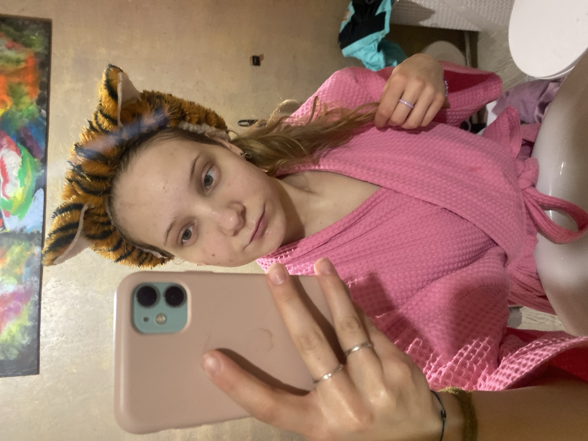 A young woman wearing a pink bathrobe and a tiger-striped headband takes a selfie with a pink smartphone. She has light skin and blonde hair. The background shows a wall, a painting, a toilet, and some clothes. The location is potentially a bathroom or bedroom in Chaikovsky, Russia.