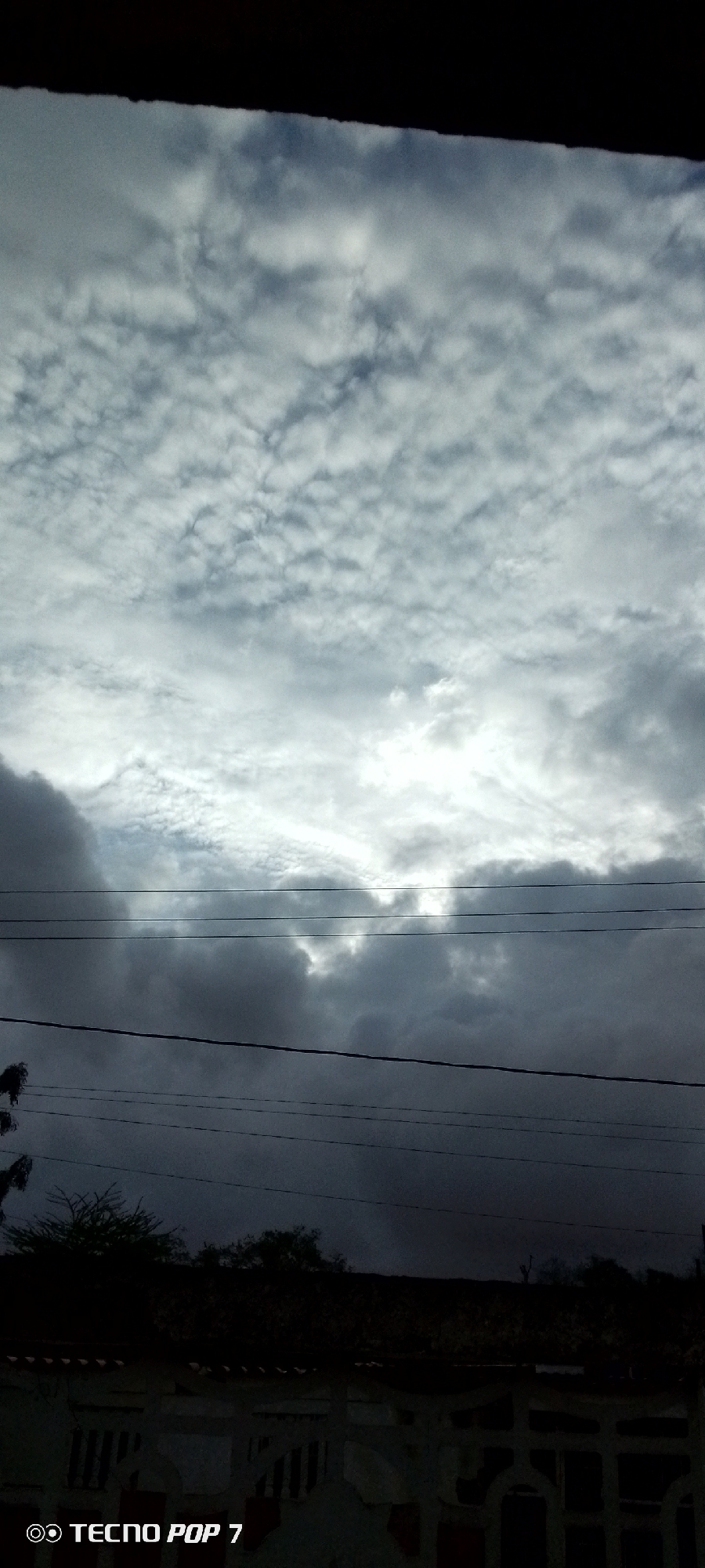 The image depicts a sky filled with a mix of light and dark clouds, suggesting a possibly overcast or stormy day. The top of the frame shows a dark horizontal band, possibly a roofline or an overhang, obscuring the very top of the sky. Below this, the clouds transition from a textured, lighter gray with many small formations to darker, heavier masses lower down.
In the middle ground, multiple horizontal wires stretch across the frame, indicating the presence of utility lines. Below the wires, silhouetted trees and the tops of structures are visible, appearing dark and indistinct against the cloudy sky. The lower part of the image reveals a white fence or railing with a patterned design.
The overall impression is one of an outdoor scene, likely in a populated area due to the power lines and structures. The lighting suggests it could be daytime, but the dense clouds create a subdued and possibly dramatic atmosphere. There is no visible activity of people.
In the bottom left corner, the text "TECNO POP 7" is visible, indicating the brand of the phone used to capture the image. The setting appears to be Altagracia, Venezuela, as provided in the location context.