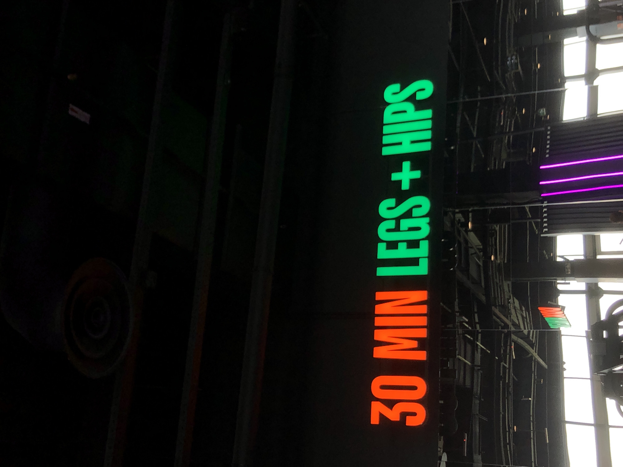 The image captures a brightly illuminated sign against a dark, industrial interior, seemingly rotated to show the text vertically. The sign prominently displays the words "30 MIN LEGS + HIPS" in large, glowing capital letters. "30 MIN" is rendered in a vibrant orange, while "LEGS + HIPS" shines in a bright green.
The background features dark structural elements and beams, suggestive of modern architecture or a gym facility. To the right of the sign, large windows allow significant natural light into the space, appearing bright white and indicating it is daytime. A vertical strip of neon purple light is also visible on the right side, adding another accent to the lighting scheme. In the upper left quadrant, a dark, circular object, possibly a vent or a speaker, is partially visible.
No individuals are present in the frame. The visible text directly points to a fitness-related activity or class, specifically a "30 minute Legs + Hips" workout. The overall setting implies a contemporary fitness environment or a sports complex, possibly located in Krasnoyarsk, Russia, given the provided context. The strong contrast between the dark background and the glowing text, along with the mix of natural and artificial lighting, creates a striking visual.