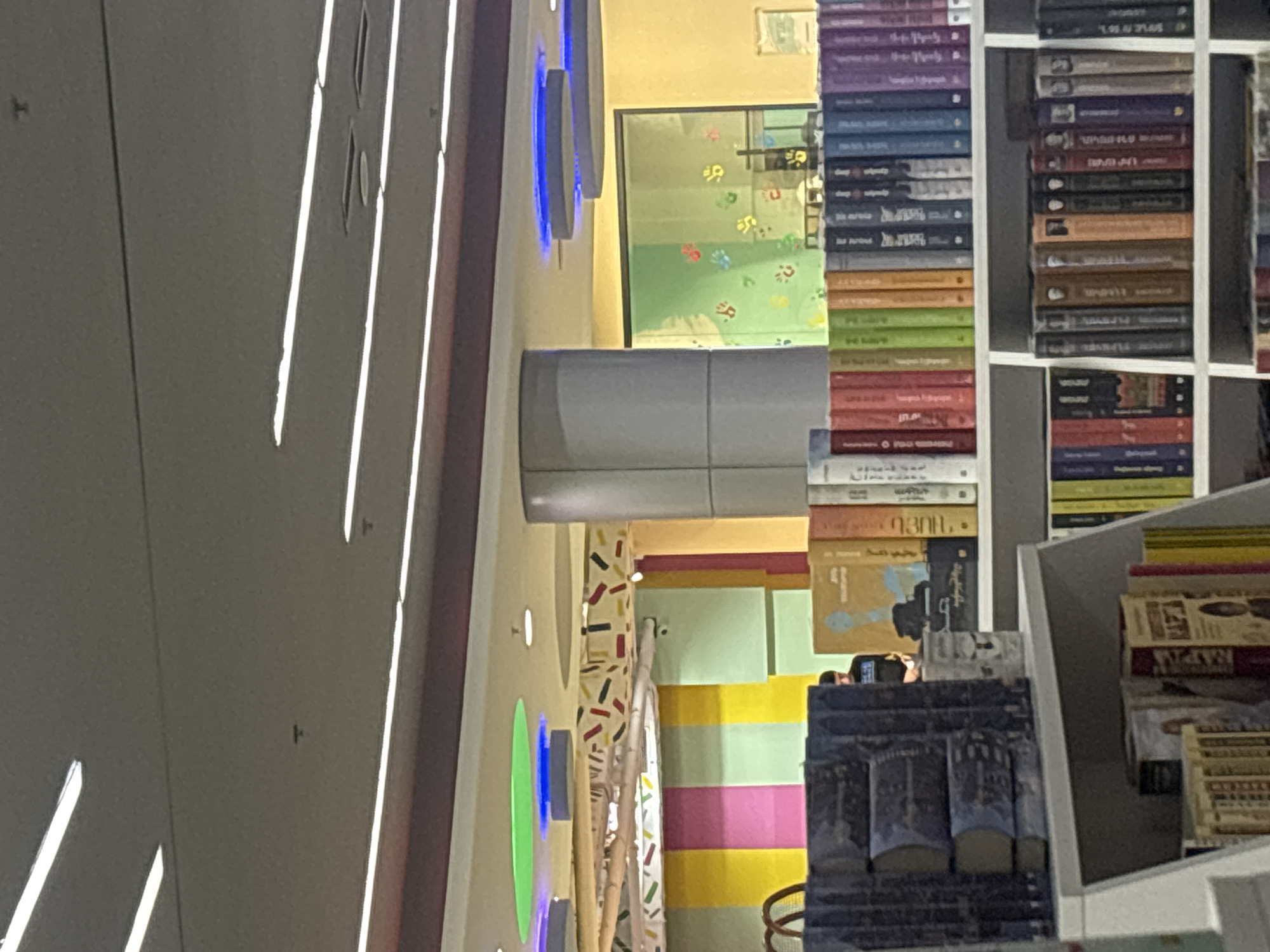 The provided image, rotated 90 degrees clockwise, captures an indoor scene strongly suggestive of a modern bookstore or a public library in Yerevan, Armenia.
On the left side of the rotated image, a gray ceiling with parallel linear light fixtures extends upwards. Adjacent to this, a cream-colored wall is adorned with several illuminated blue circular and green oval light installations. A large, reflective silver cylindrical pillar stands centrally. Below this, a glass panel or window displays multiple colorful paw prints in red, green, blue, and yellow, set against a light green background, indicating a child-friendly or thematic area. A gray padded bench is partially visible beneath this window.
The right side of the image is filled with tall, white bookshelves, densely packed with books stacked vertically. The book spines exhibit a wide spectrum of colors, including purple, blue, green, red, brown, and black. Although much of the text is indistinct, several book spines clearly display text in the Armenian alphabet, confirming the location context of Yerevan, Armenia. The word "ԱՐՄԵՆԻԱ" (Armenia) is notably visible on one book. Some spines also appear to feature short English words like "deep" and "sleep," suggesting a mix of languages or translated works.
A person is partially visible in the lower right section of the image, largely obscured by shelves and books, appearing to browse or use a mobile phone. In the background, behind the lower bookshelves, a vibrant, multi-colored wall with horizontal stripes in shades of light green, yellow, and pink adds to the lively atmosphere. Another decorated wall segment features a pattern of small, scattered rectangular shapes in red, black, and yellow.
The setting is brightly lit, showcasing a modern and engaging environment designed for reading and cultural engagement.