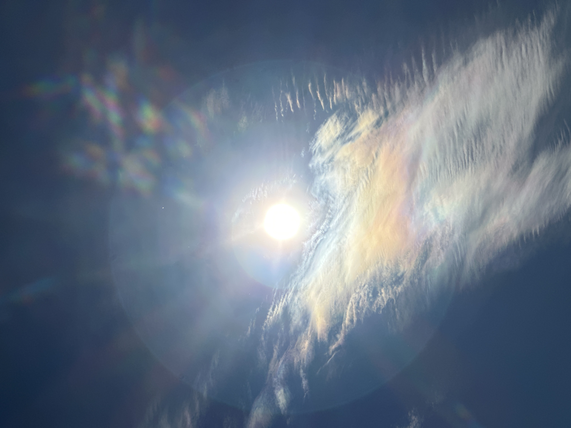 The image depicts a bright sun shining through clouds, creating a lens flare effect. There is cloud iridescence, a rainbow-like coloration, in the clouds to the right of the sun, as well as on the left side of the sun, creating a prism-like effect. The environment appears to be outdoors. There is no indication of the location being Nizhny Novgorod, Russia, or any specific city. There are no people or interactions visible in the image.