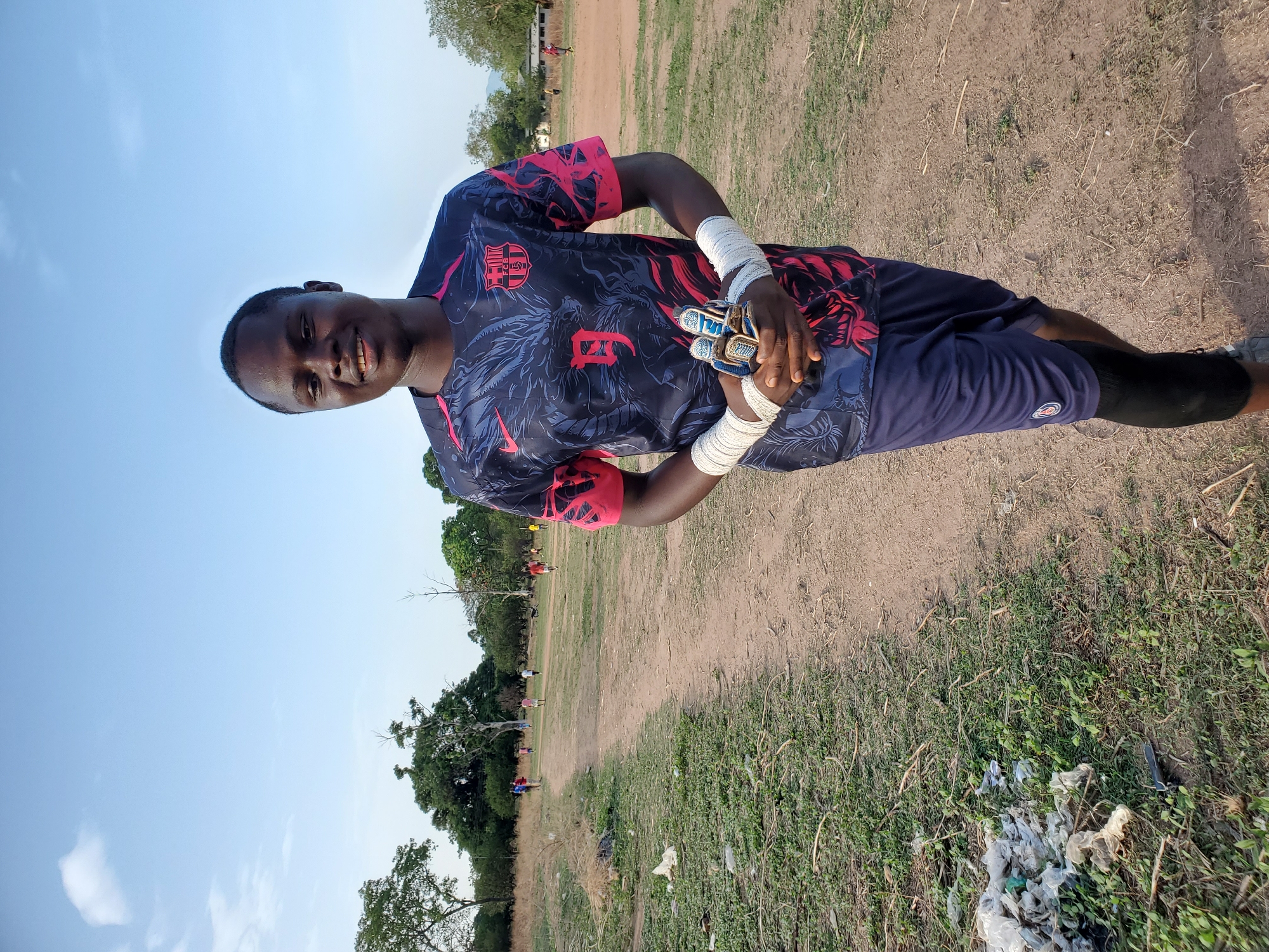 An outdoor, portrait-oriented shot captures a young man smiling at the camera on a sports field in Mararaba, Nigeria. He is positioned centrally, wearing a dark blue and red patterned FC Barcelona football jersey with a prominent Nike logo. A large, stylized letter or number resembling a "B" or "9" is also visible on the front of his shirt. He has white athletic wraps on both wrists and is holding a pair of blue and white goalkeeper gloves, one of which clearly displays "2000" on a finger. He is also wearing dark blue shorts over what appear to be black compression shorts or leggings.
The background reveals a dry, unpaved dirt and grass field, characteristic of a local sports pitch. Scattered trees line the far edge of the field, and several distant figures of other people can be seen, suggesting an ongoing or upcoming activity like a football match. The sky is clear and light blue, indicating daylight, possibly late afternoon given the soft lighting. In the foreground to the right, some litter, mainly white plastic bags, is visible on the ground amidst sparse vegetation. The young man's expression is bright and positive.