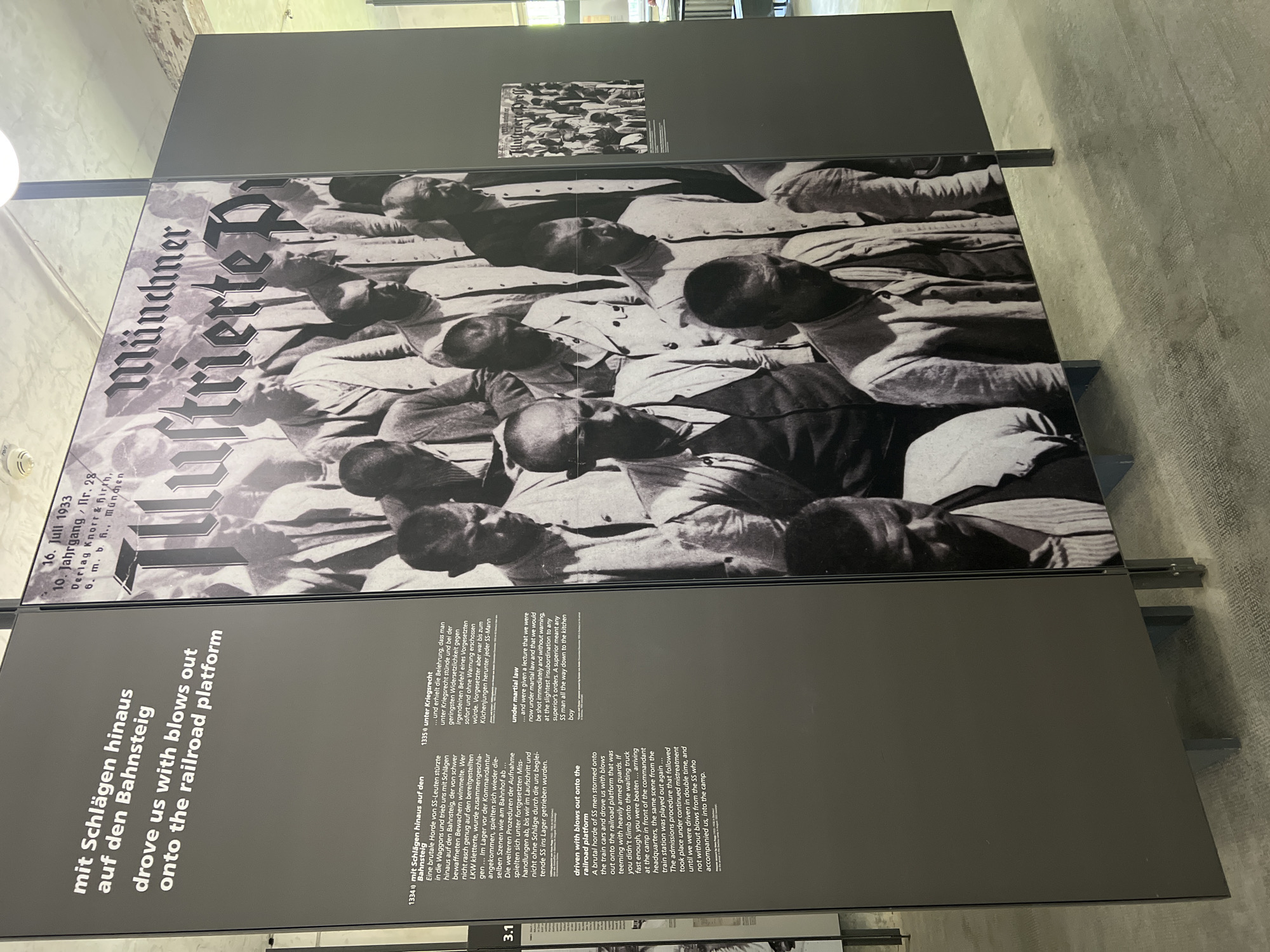 The image displays an exhibit at the Dachau Concentration Camp Memorial Site. The exhibit features a large black and white photographic mural depicting a group of men, likely prisoners, wearing civilian clothing. The mural is accompanied by text panels in German and English.
The English text, titled "driven us with blows out onto the railroad platform," describes a brutal experience of being forced out of wagons onto a railroad platform by SS men. It recounts the chaotic arrival at the camp, the intimidating presence of armed guards, and the subsequent admissions procedure.
To the right of the mural, a smaller black and white photograph shows a group of men, possibly the same individuals or similar in context, with a newspaper headline visible above them that reads "Münchner Illustrierte Presse," dated "16. Juli 1933." This suggests the larger mural might be a reproduction or an artistic interpretation related to historical documentation from that period.
The overall environment appears to be an indoor exhibition space, with modern display panels and lighting. The floor is a patterned carpet. The display is designed to convey historical information and personal experiences from the Dachau concentration camp.