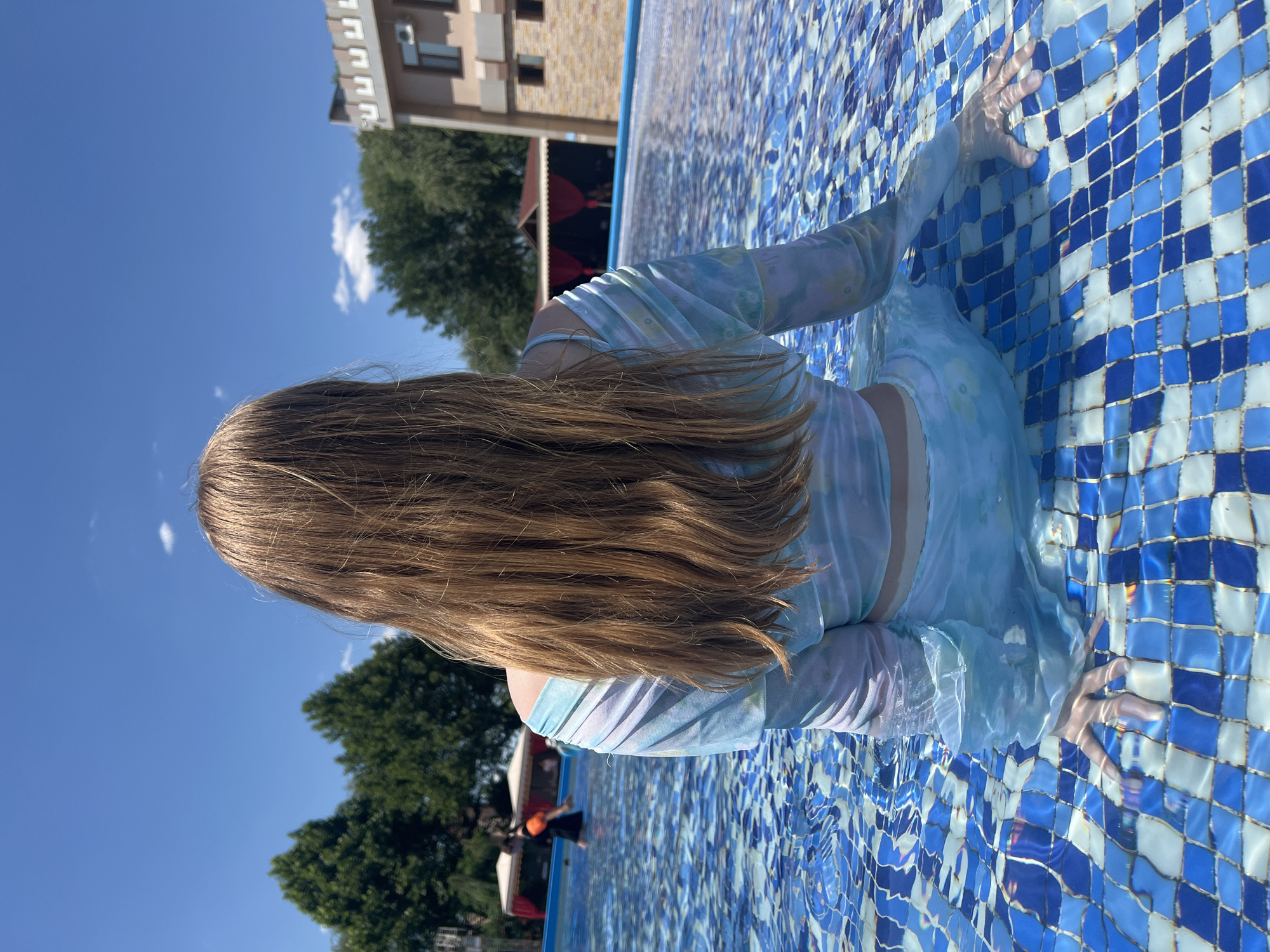 A person with long, blonde hair is sitting in a swimming pool, facing away from the camera. They are wearing a light blue and white tie-dye swimsuit. The pool has a blue and white checkered tile bottom. In the background, there are trees, a building with a textured facade, and a red awning. The sky is clear blue with a few white clouds. There are other people visible in the pool in the distance. The image appears to have been taken on a sunny day.