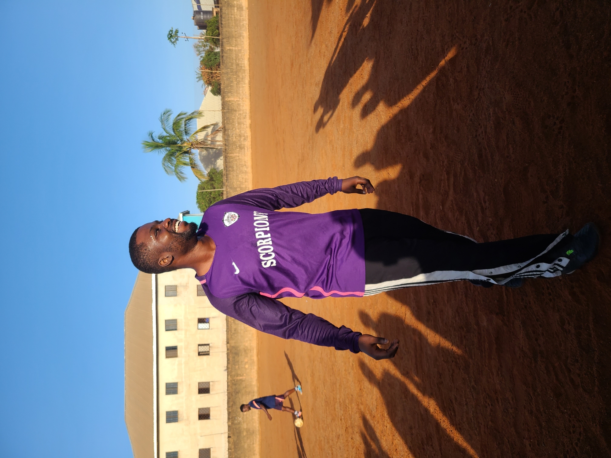 An outdoor shot depicts a man smiling broadly and looking upwards, standing on a reddish-brown, unpaved surface, likely a sports field or dusty ground. He wears a purple long-sleeved athletic shirt with the word "SCORPION" in white text and a white Nike swoosh logo. His pants are dark with white stripes along the sides, and he wears dark athletic shoes.
In the background, to the left, a smaller figure is visible, appearing to be a person (possibly a youth) in a dark shirt and shorts, standing near a soccer ball. Long, dark shadows stretch across the ground from multiple sources, indicating that the sun is low in the sky, suggesting either early morning or late afternoon. The sky is clear and bright blue.
Behind the field, a tall, multi-story building with numerous windows is visible, along with a concrete block wall and some sparse vegetation, including a prominent palm tree. The overall setting suggests a warm, sunny environment. The main subject appears to be happy and engaged, possibly taking a break or posing during an activity.