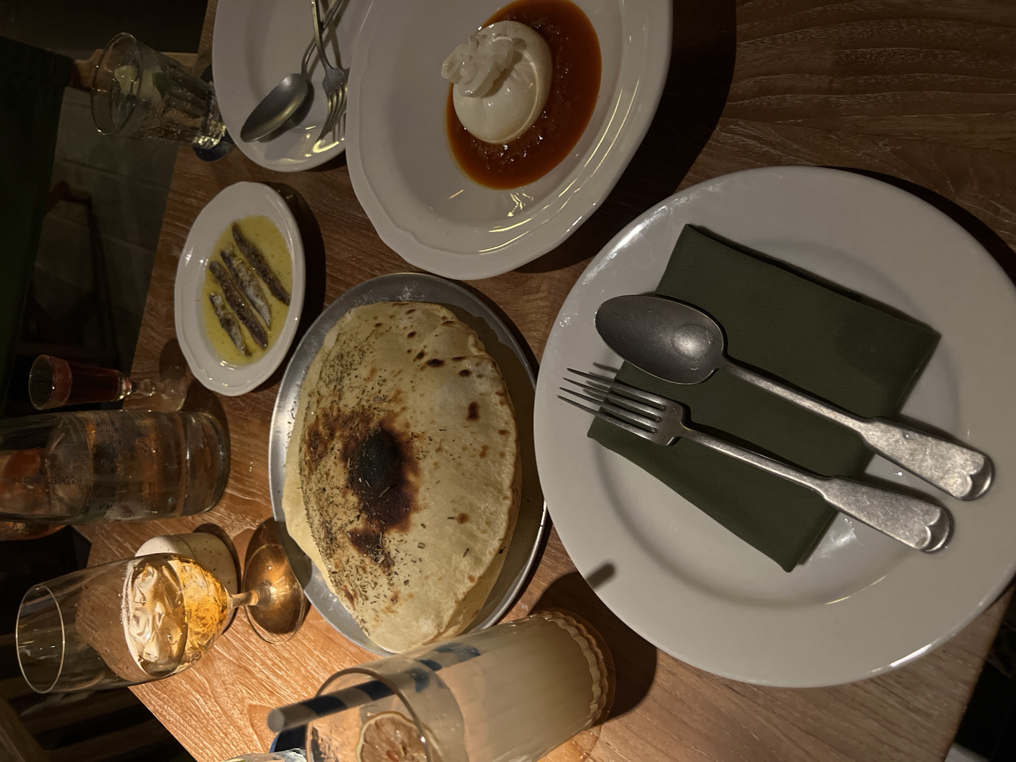 On a wooden table in North Kuta, Indonesia, a meal is laid out. The scene includes a glass of white wine on a coaster, a carafe of water, and a small glass with a reddish liquid. A plate of anchovies in oil sits near a glass of iced beverage with a straw and a slice of lemon. A large, round flatbread is presented on a metal tray. Further to the right, a white bowl contains a ball of burrata cheese in a red sauce. In the foreground, a white plate holds a green napkin, a fork, and a spoon. The lighting suggests an indoor setting, possibly a restaurant.