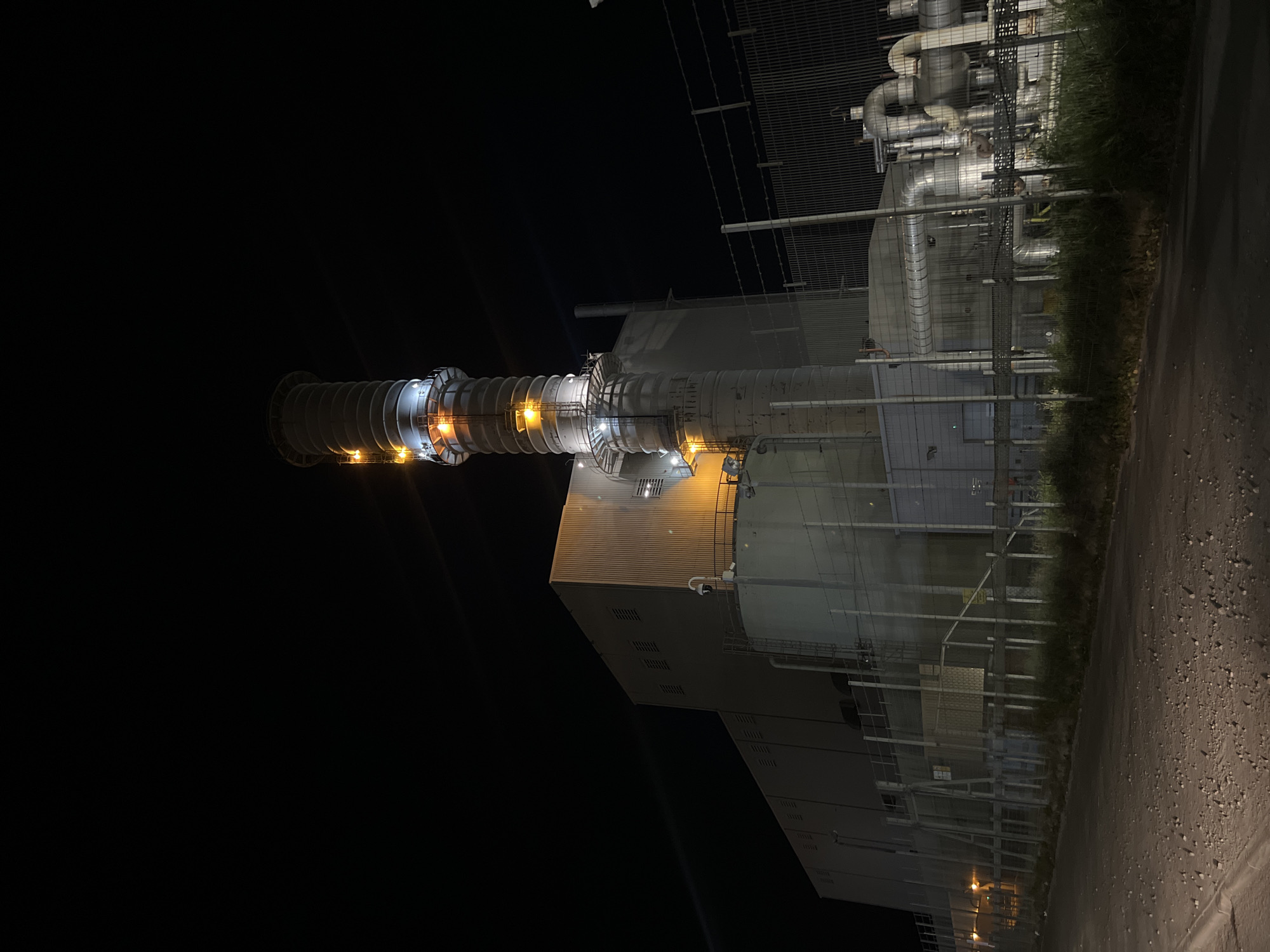 The image depicts a nighttime view of what appears to be an industrial facility in Gorleston-on-Sea, United Kingdom. The main subject is a large structure with cylindrical components and a building-like section illuminated by several lights. The structure is surrounded by fencing, pipes, and vegetation. The ground is dark and textured, possibly asphalt or gravel. The background is primarily black, indicating a night scene. There are no discernible people or specific actions visible.