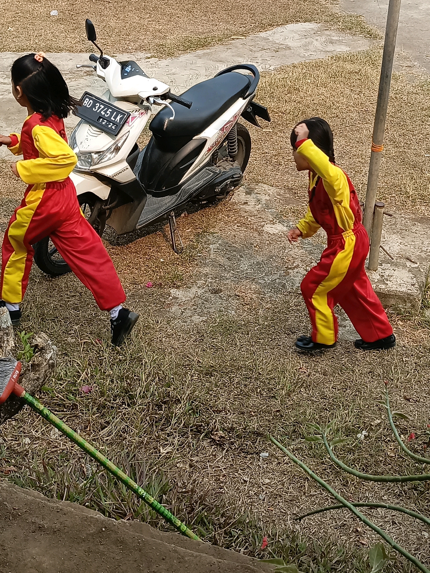 Two young children, dressed in matching red and yellow jumpsuits, are on a dirt and grass-covered ground next to a white motorcycle. The child on the left, facing away from the camera, is walking towards the left side of the frame. The child on the right, facing left, is in motion, with one hand raised to their head. The motorcycle, with a license plate reading "BD 3745 LK 12-24", is parked on the ground. A wooden pole stands to the right of the children, and a broom is visible in the lower left corner. The environment suggests an outdoor setting, possibly a yard or a public space in Taba Tinggi, Indonesia, indicated by the presence of the motorcycle and the style of the license plate.