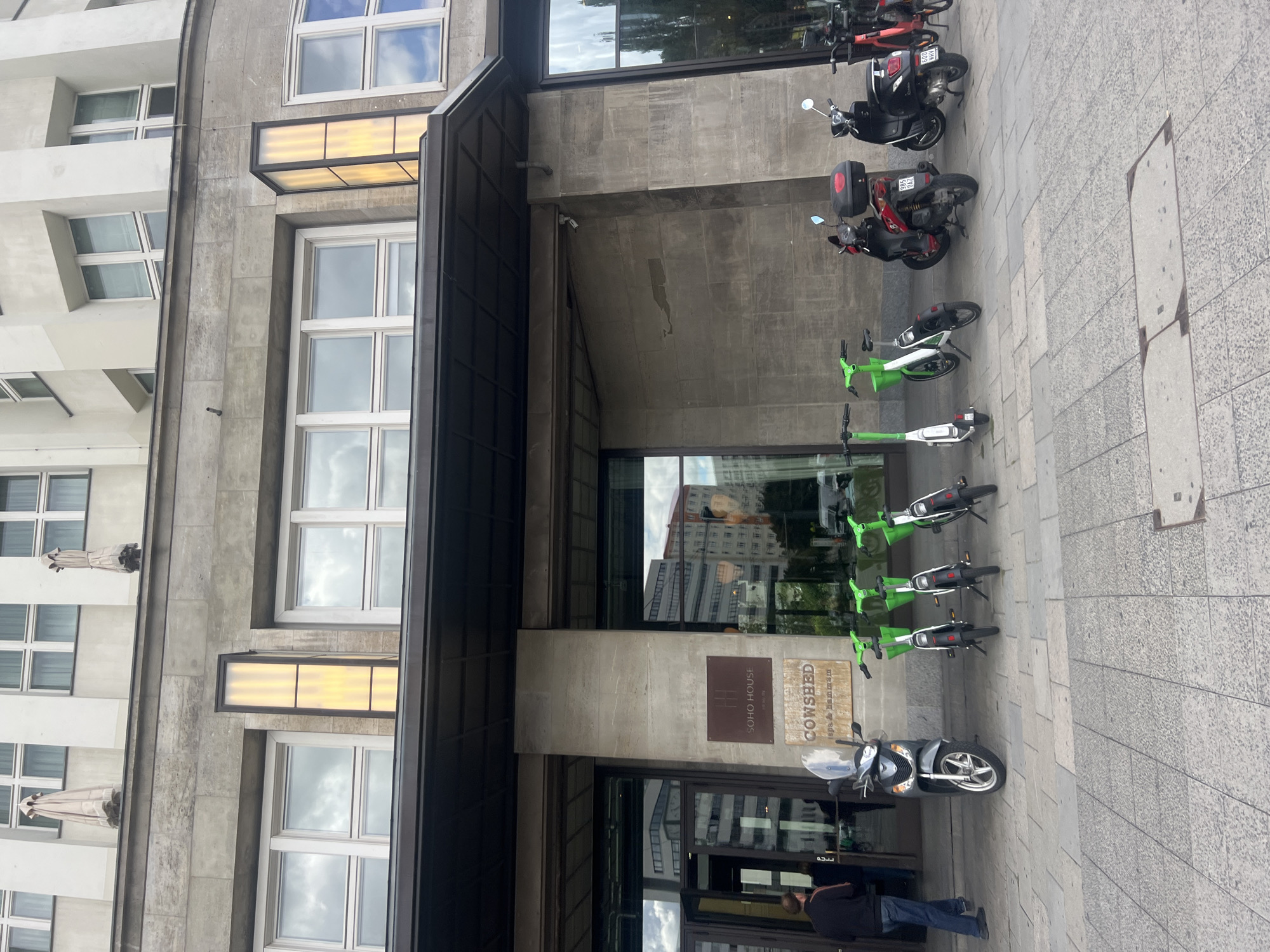 The image depicts the entrance to Soho House in Berlin, Germany. The building features a brutalist architectural style with large concrete blocks and prominent windows. In front of the entrance, there is a prominent Soho House sign and a Cowshed spa sign. Several lime-green electric scooters are parked to the right of the entrance, along with three dark-colored motor scooters and one red motor scooter parked further to the right. A person in a dark jacket and blue jeans is walking away from the entrance towards the left side of the frame. The pavement is made of large square tiles. The sky is cloudy, with glimpses of blue sky and reflections of buildings in the windows.