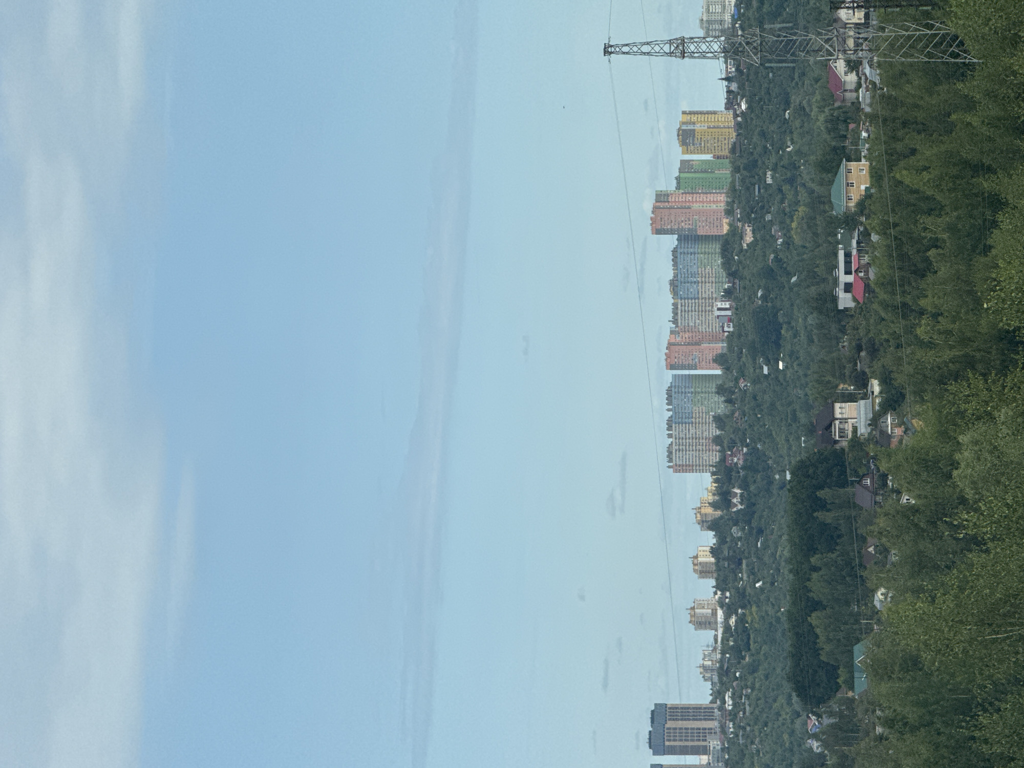 The image captures a panoramic view of a suburban landscape transitioning into a city skyline, likely in Ankudinovka, Russia. In the foreground, dense green trees create a natural barrier, with scattered houses and smaller buildings visible amongst them. The middle ground features a more developed residential area with various houses and a prominent power line tower on the right side. The background is dominated by a modern city skyline with several tall, multi-colored apartment buildings, suggesting a significant urban development on the horizon. The sky is a pale blue with scattered clouds, indicating a clear day. There are no people visible in the image.