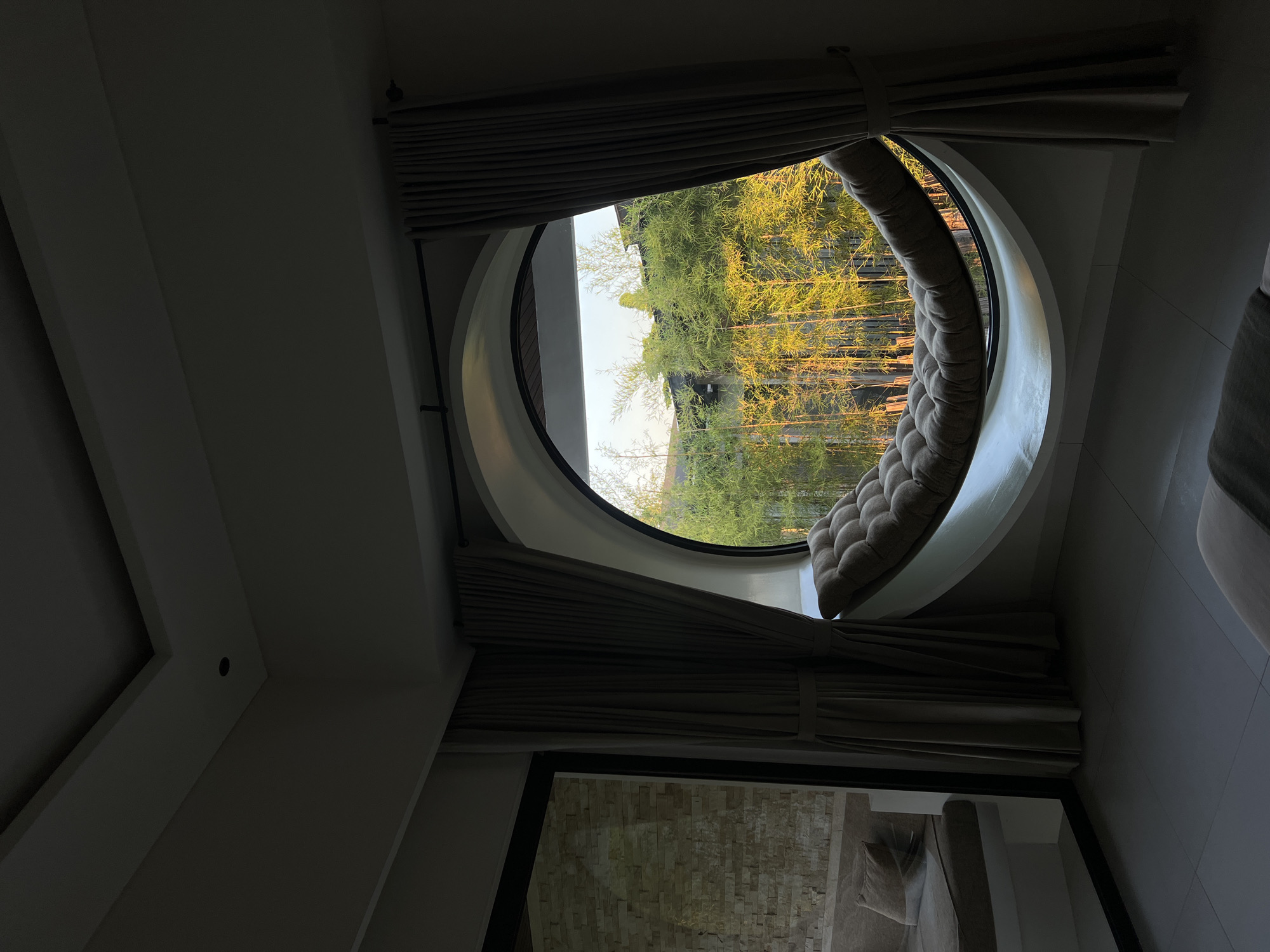 The image shows an interior view of a room with a large, circular window overlooking a bamboo forest. The window has a built-in padded seat that follows the curve of the glass. The room is dimly lit, with light streaming in from the window, illuminating the bamboo trees outside. To the left of the window, there is a glass door that reflects a stone wall and a sofa. The floor is tiled, and there is another sofa visible in the foreground. The overall ambiance is serene and natural, suggesting a tranquil living space. The location is identified as the Ubud District in Indonesia.